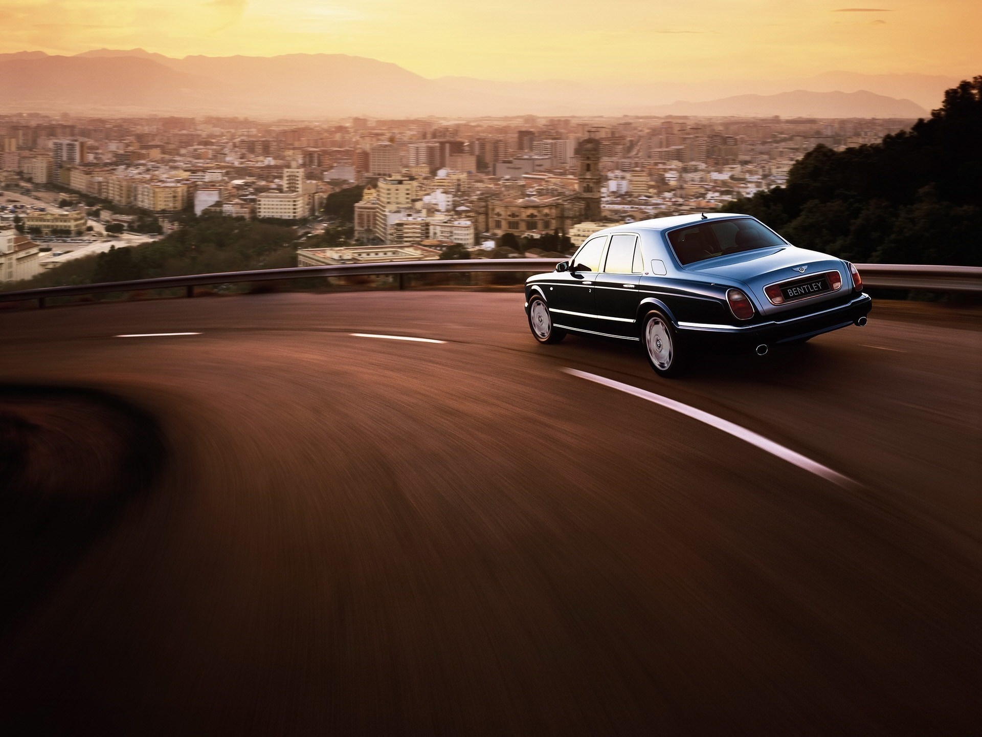 Bentley driving on a winding road overlooking a city at sunset HD Wallpaper