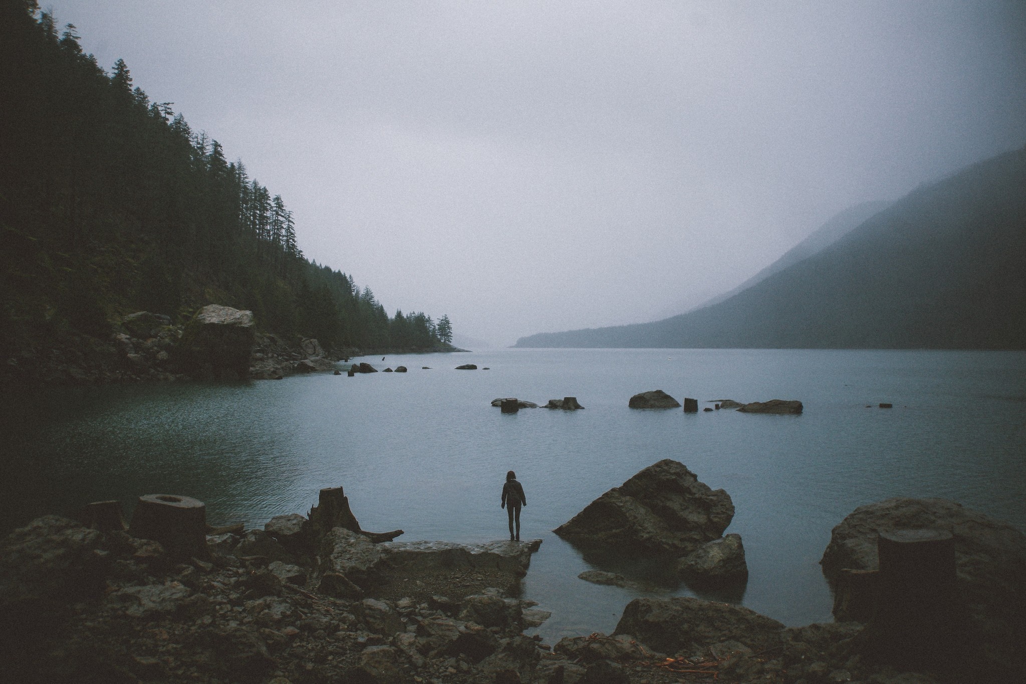 Person standing on rocks by a misty lake with mountains HD Wallpaper