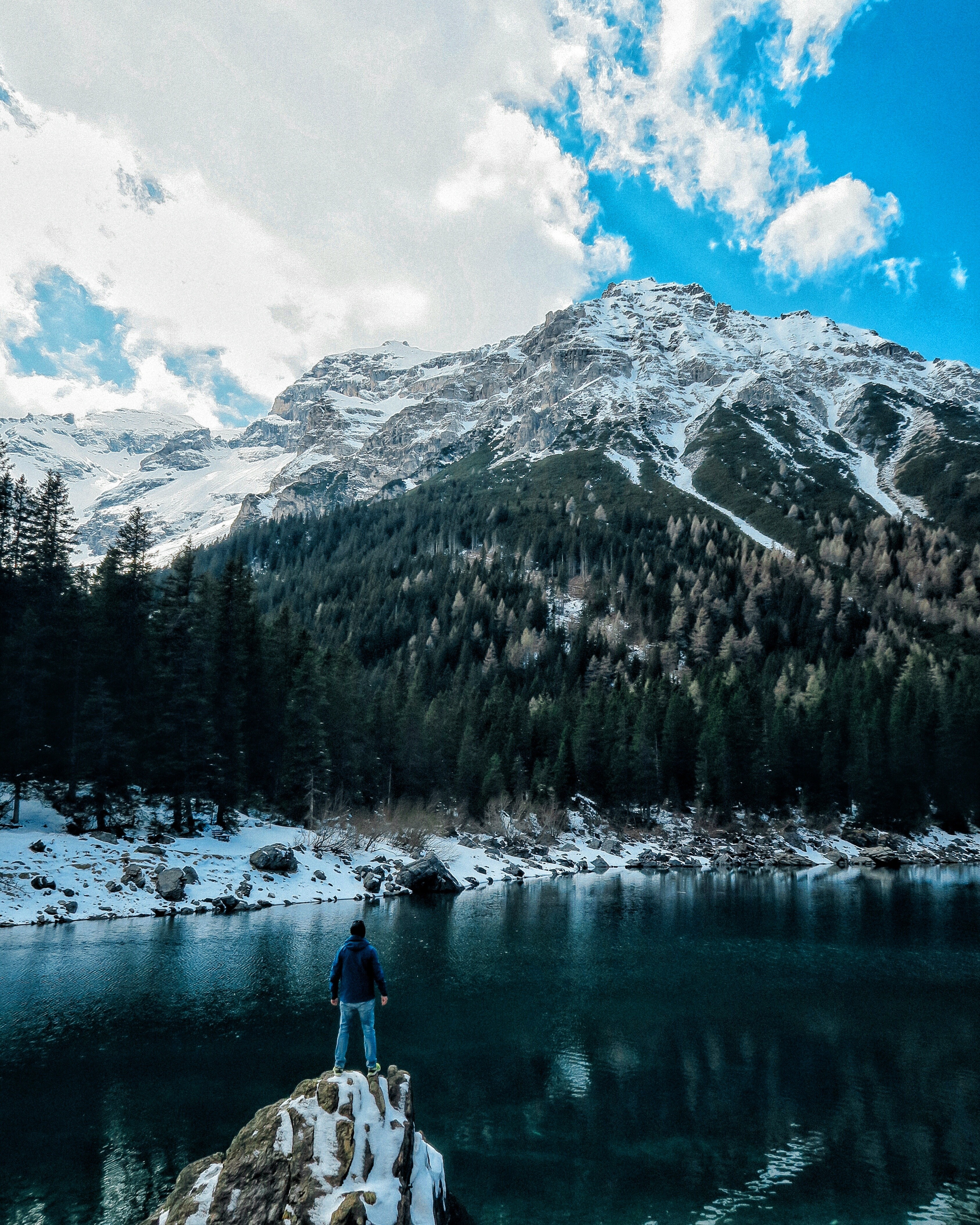 Man standing on rock overlooking snowy mountain lake HD Wallpaper