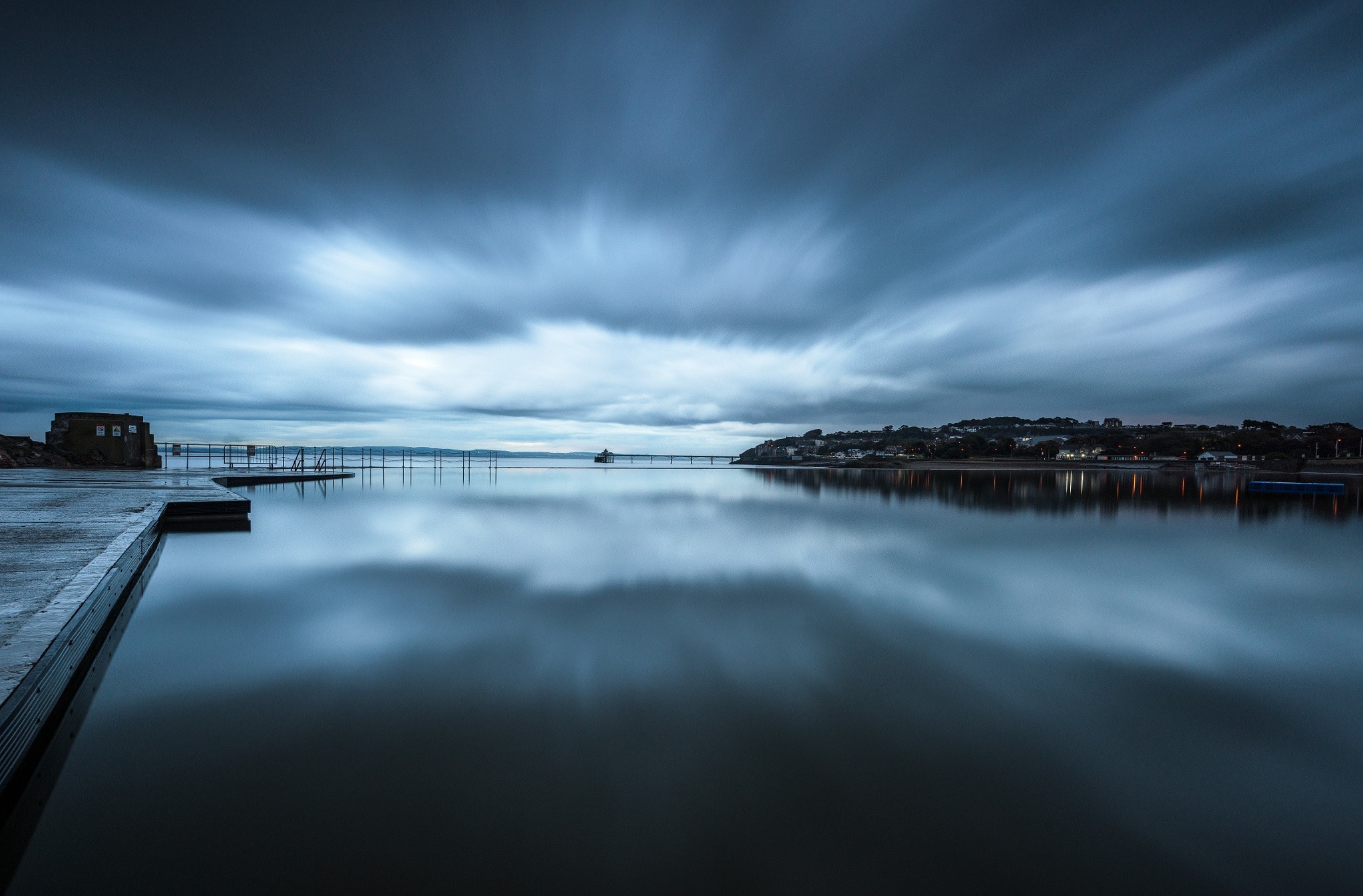 Long exposure of a calm sea with a pier and distant town HD Wallpaper