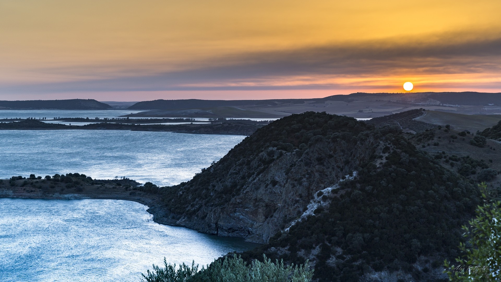 Sunset over a vast body of water with rolling hills HD Wallpaper