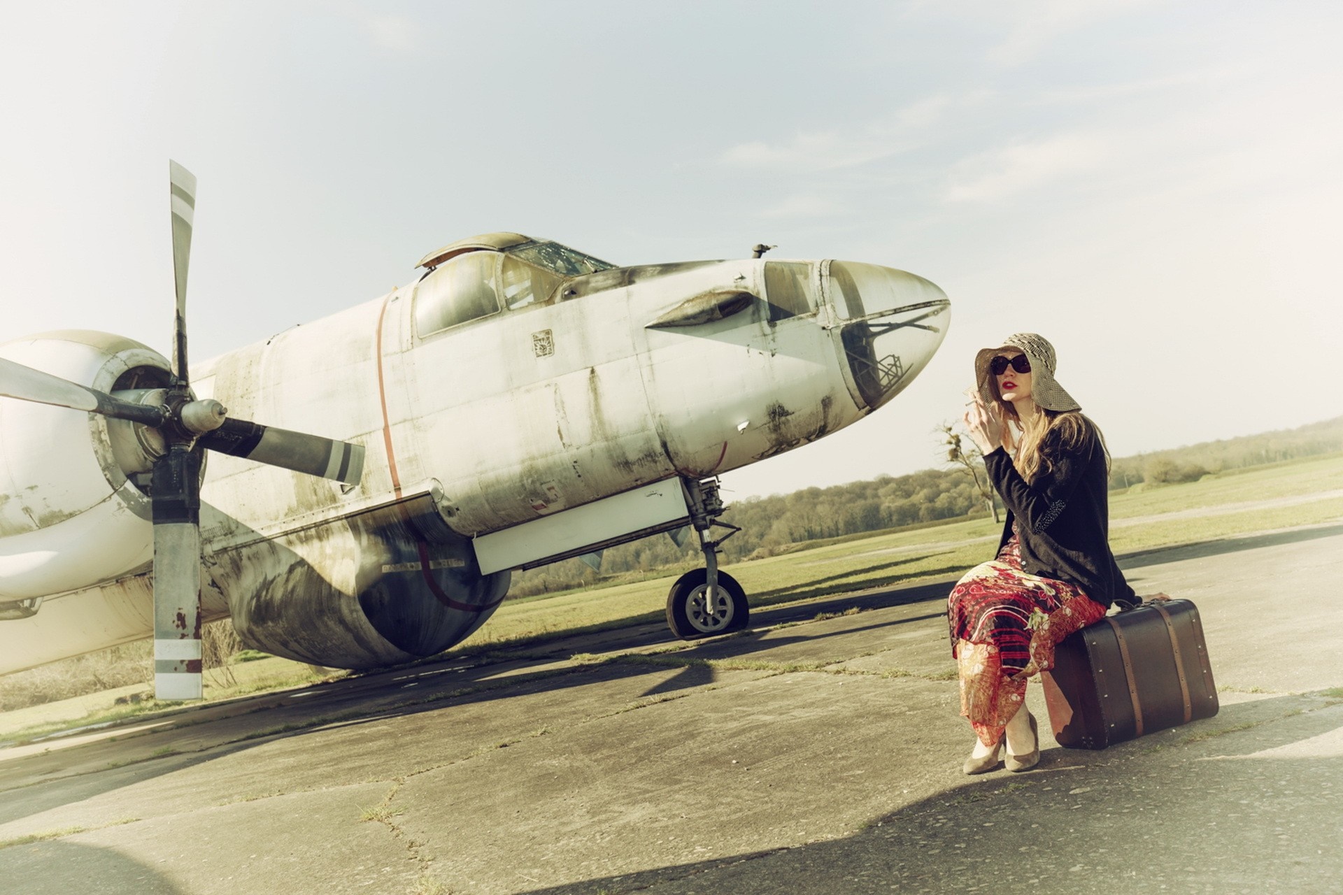 Woman in hat and sunglasses sitting on suitcase by old airplane HD Wallpaper