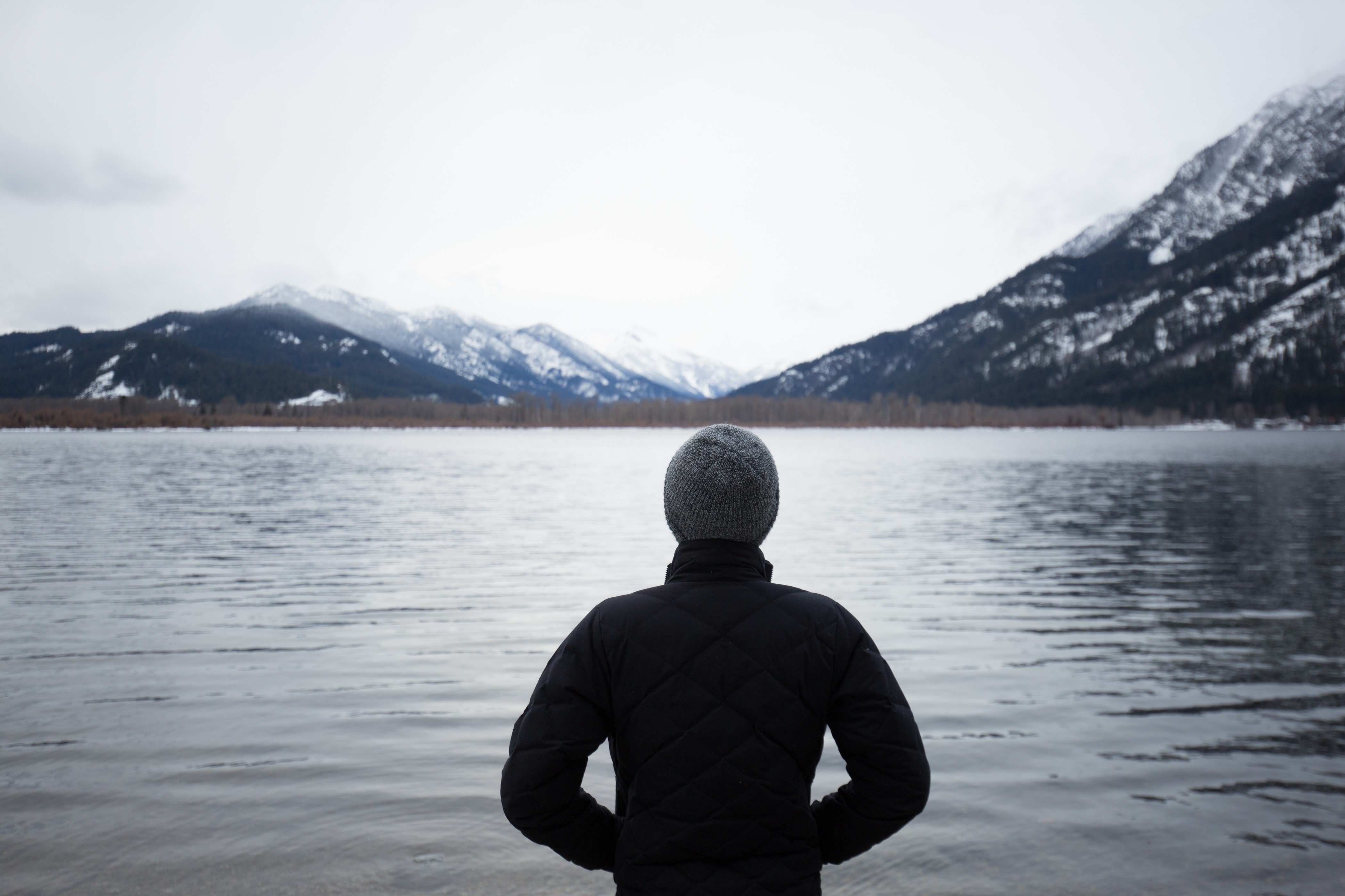 Person in a hat and jacket by a lake with mountains HD Wallpaper