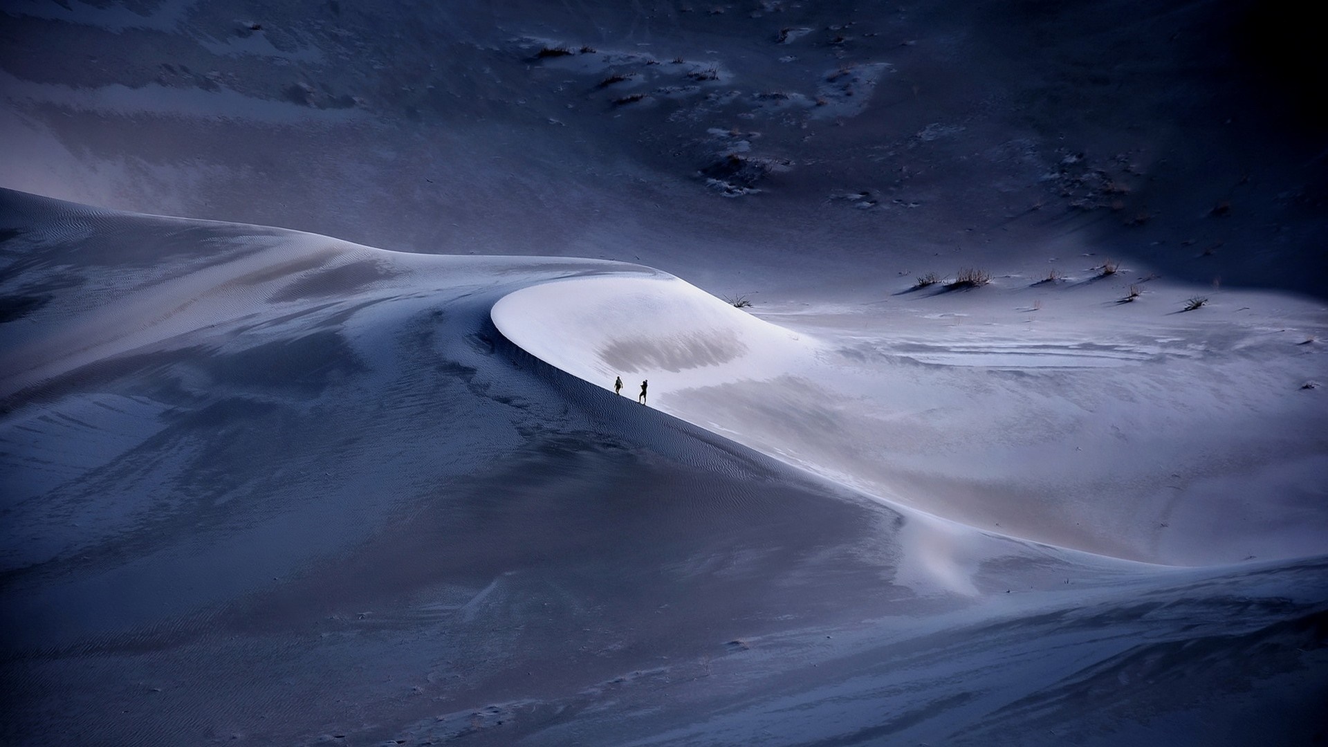 Two people walk on a sand dune in a cold, blue landscape. HD Wallpaper