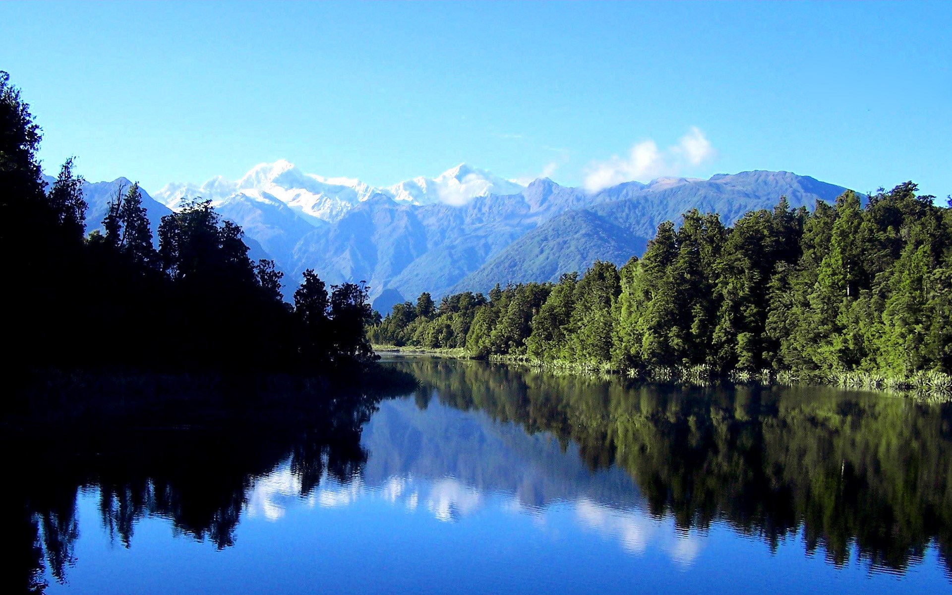 Snow-capped mountains reflected in a calm lake with evergreen trees HD Wallpaper