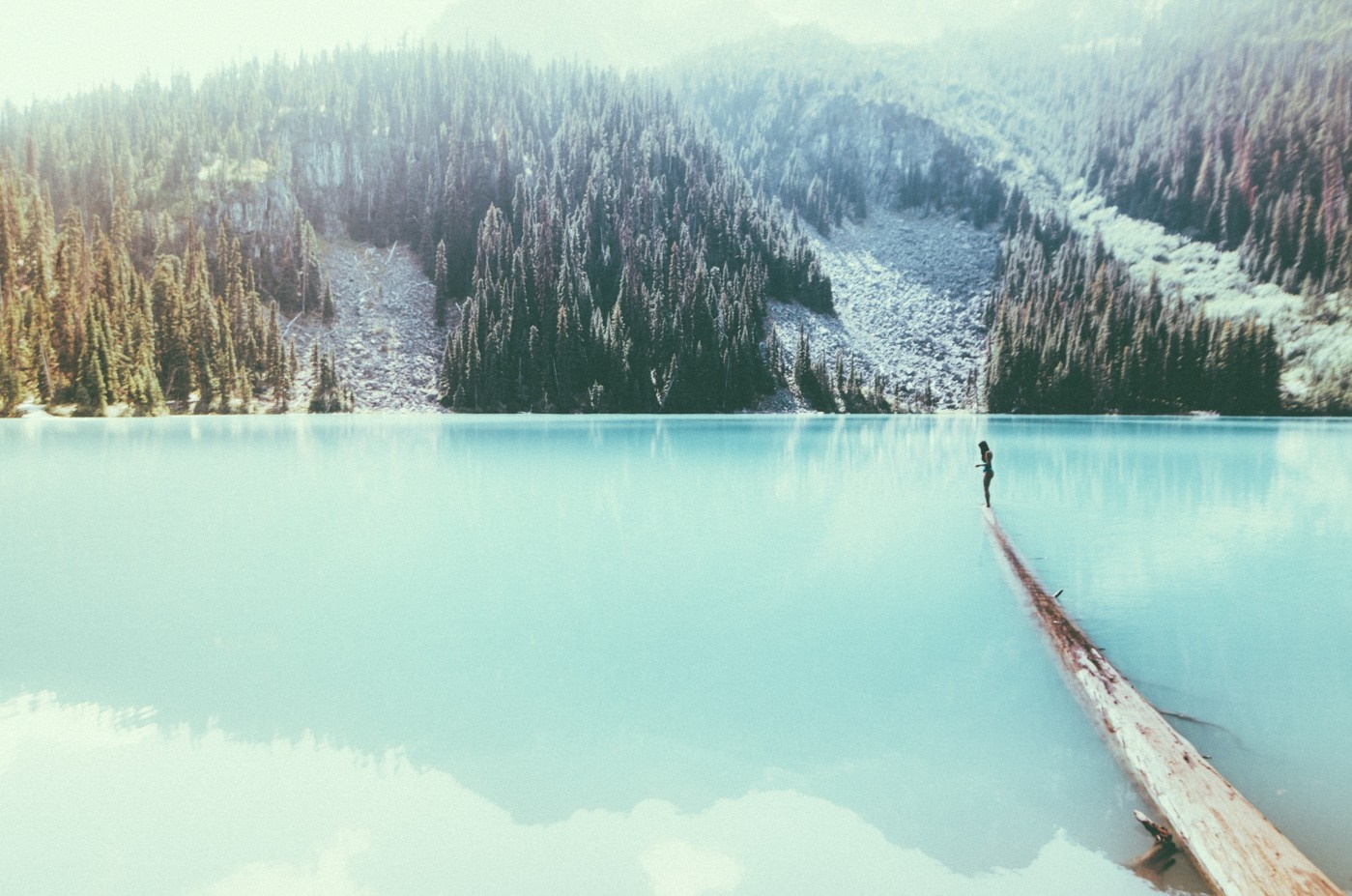 Woman on a log in a turquoise lake with mountains HD Wallpaper