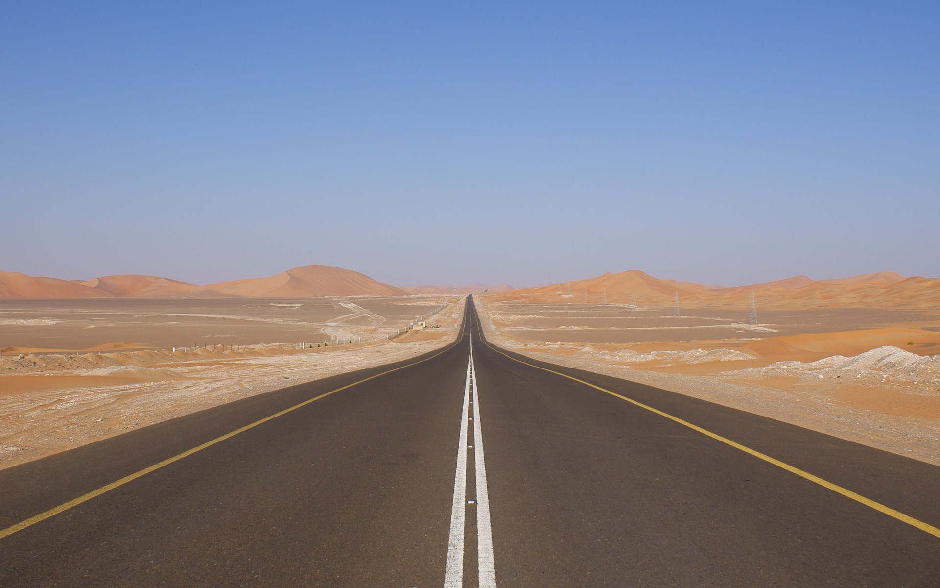 Desert highway with sand dunes under a clear blue sky HD Wallpaper