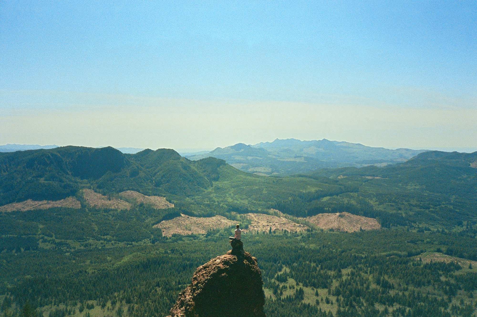 Person sitting on a rocky summit overlooking a vast forested valley HD Wallpaper