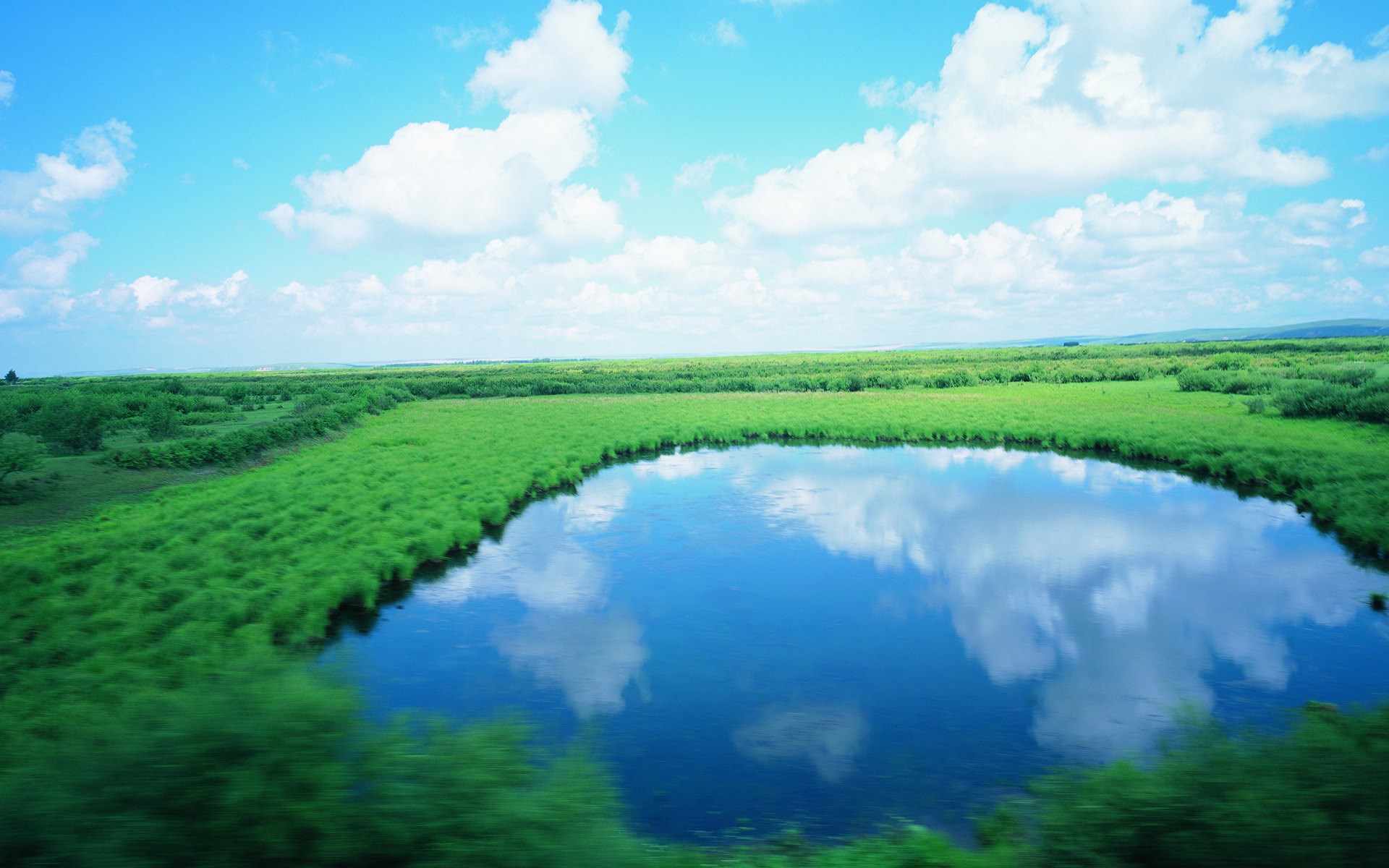 A tranquil lake reflects the sky and clouds amidst green fields. HD Wallpaper