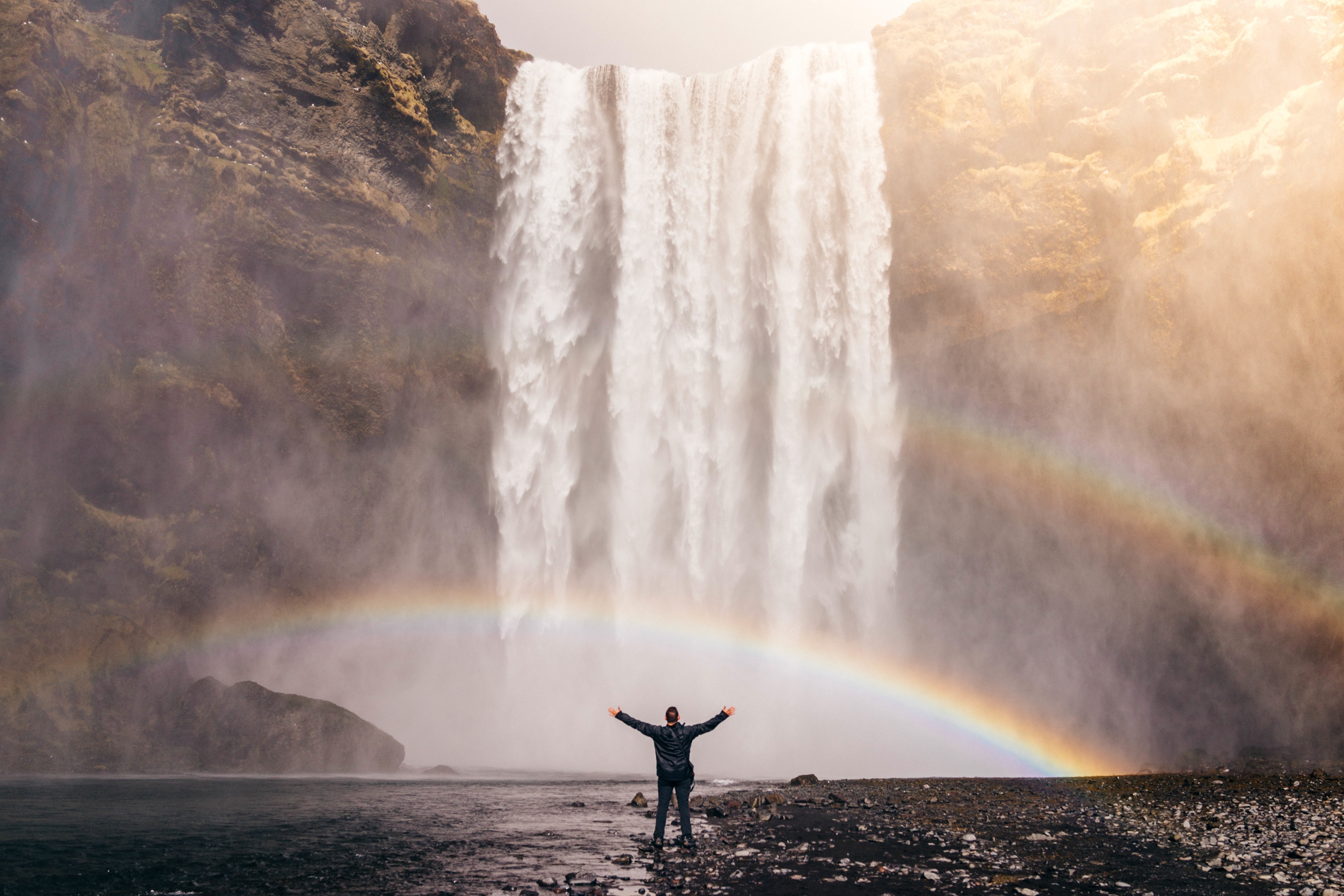 Man with arms outstretched in front of Skogafoss waterfall with rainbow HD Wallpaper