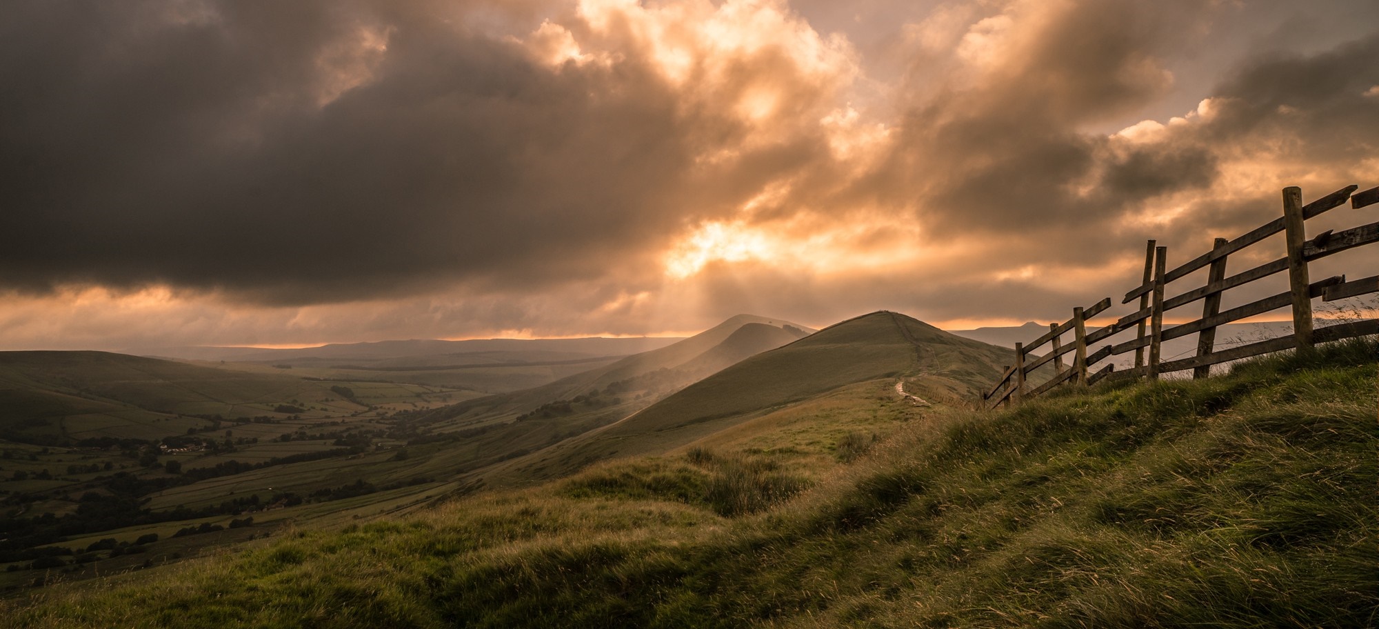 Sunlight breaking through clouds over rolling hills and a fence HD Wallpaper