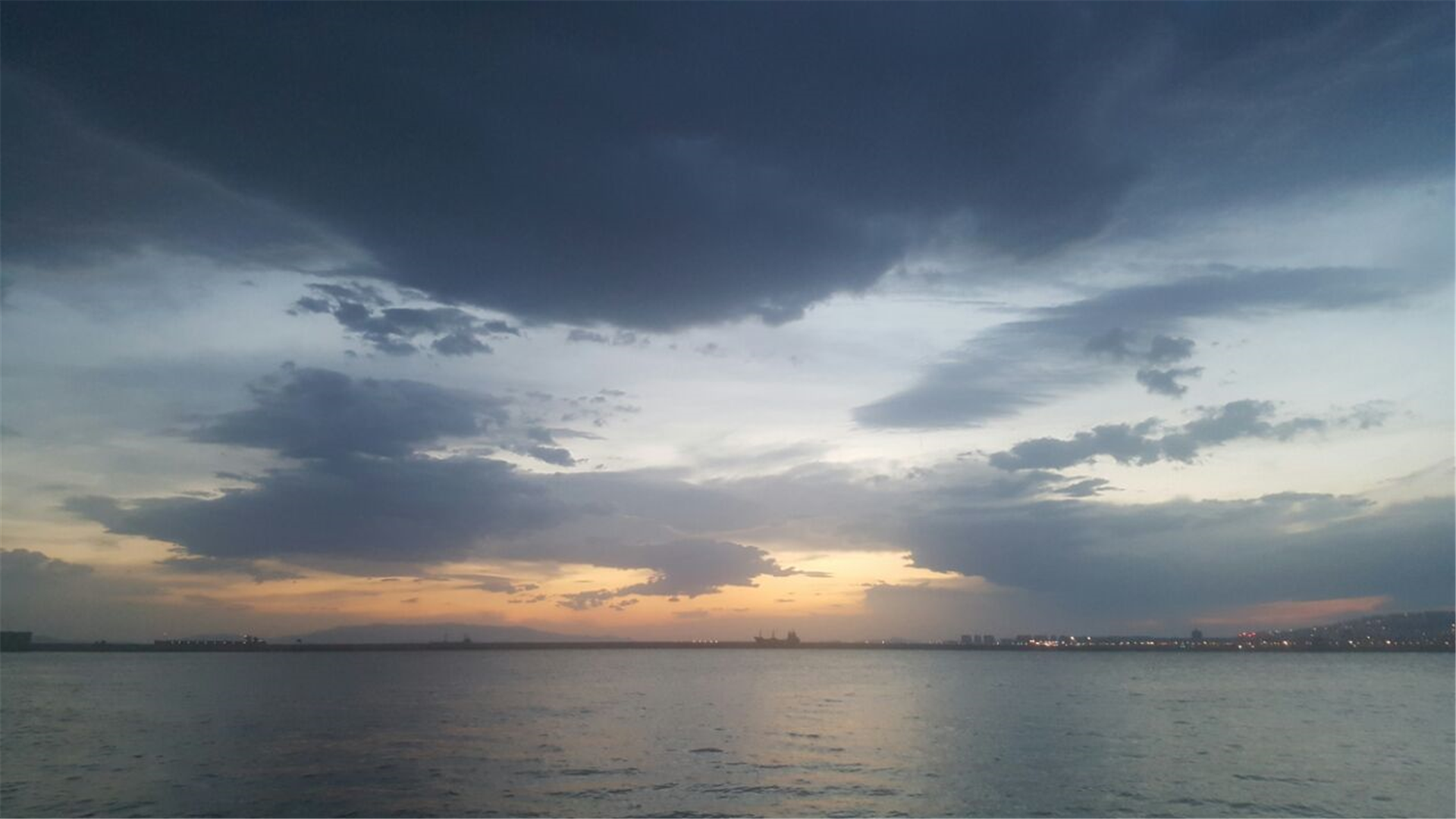 Sunset over the sea with city lights on the horizon HD Wallpaper