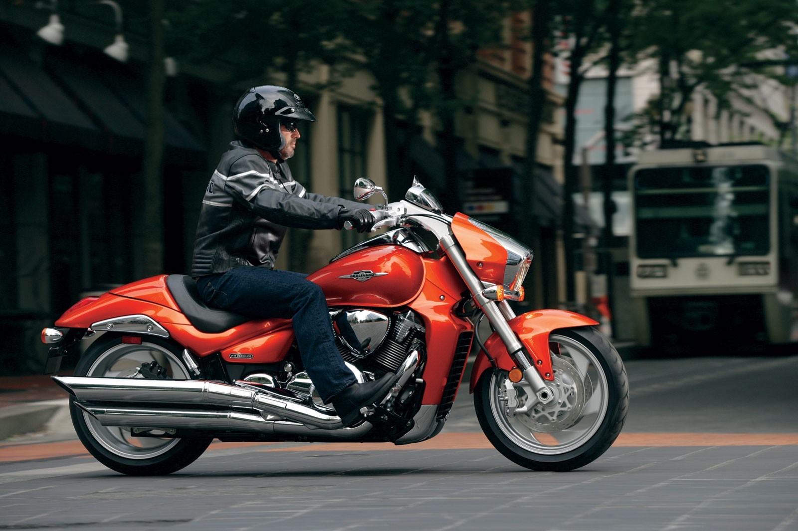 Man riding an orange Suzuki motorcycle on a city street HD Wallpaper