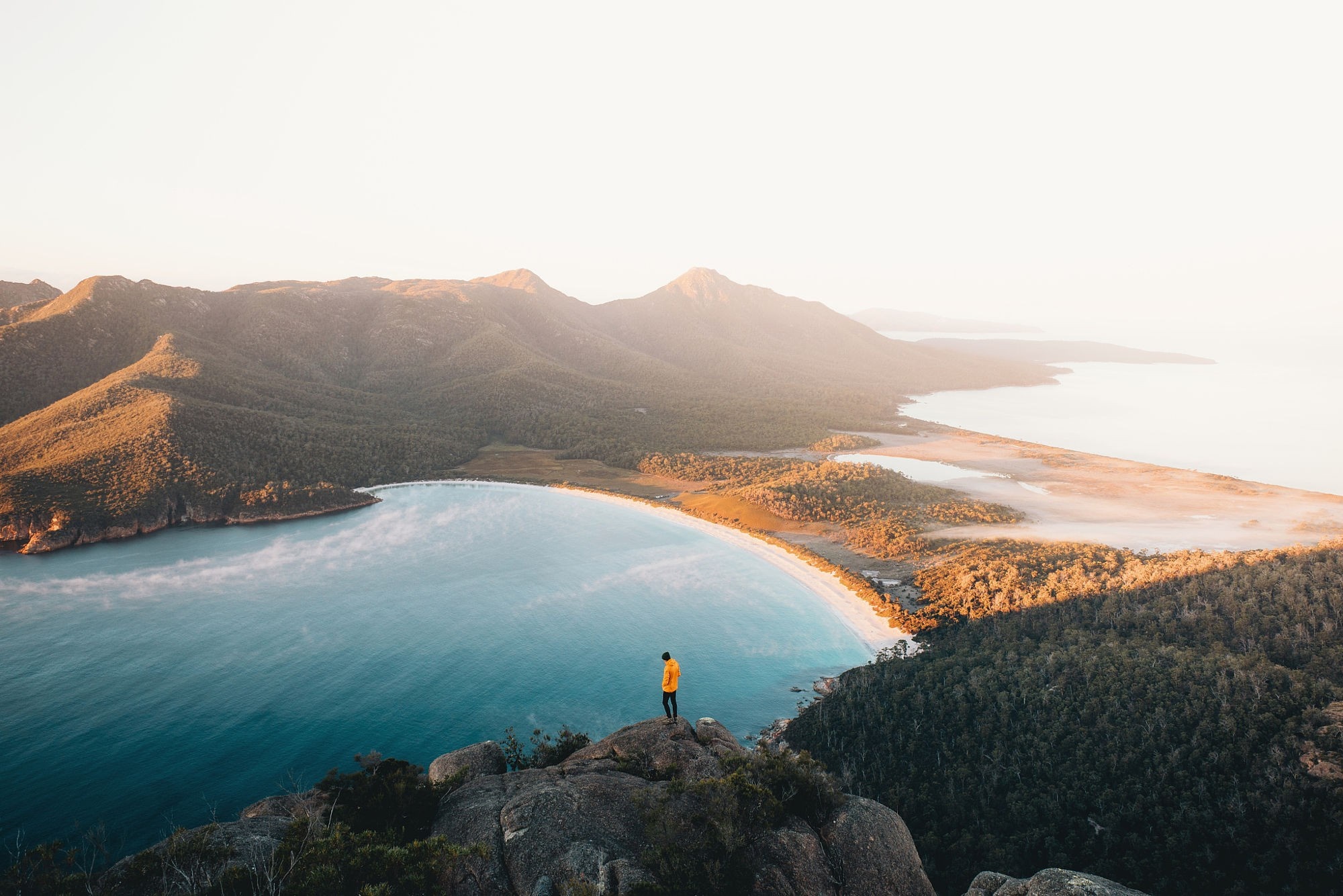 Person in yellow jacket on cliff overlooking bay and mountains HD Wallpaper