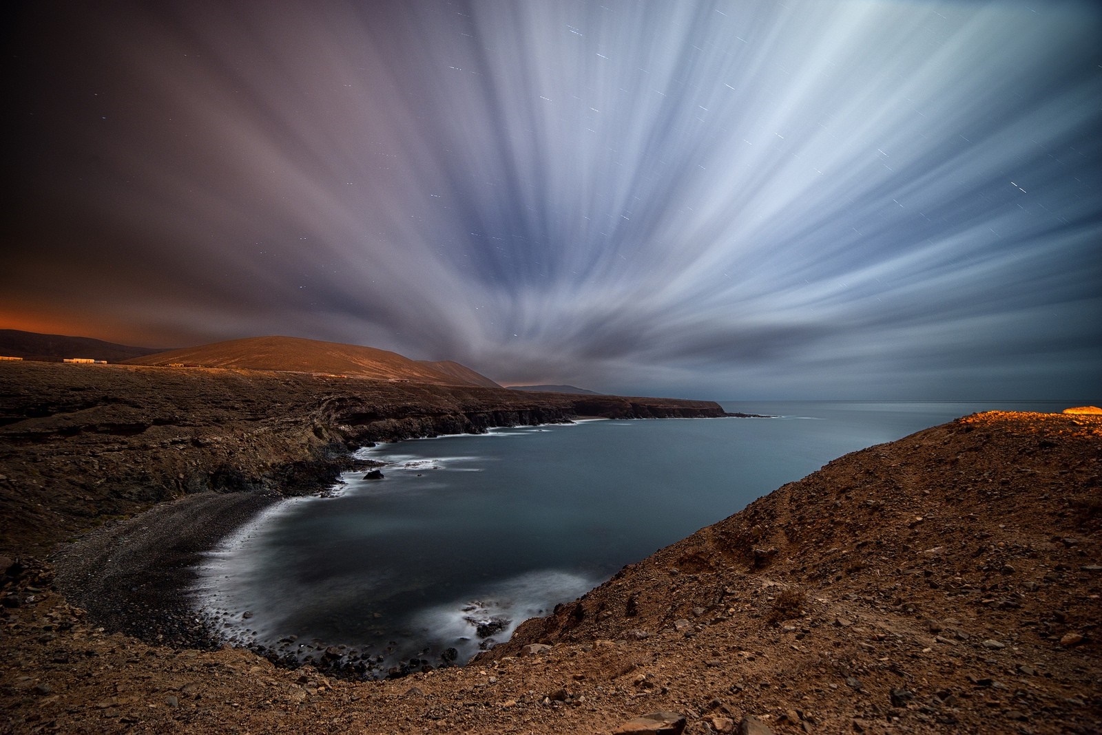 Long exposure of a rocky coast at dusk with light trails in the sky HD Wallpaper