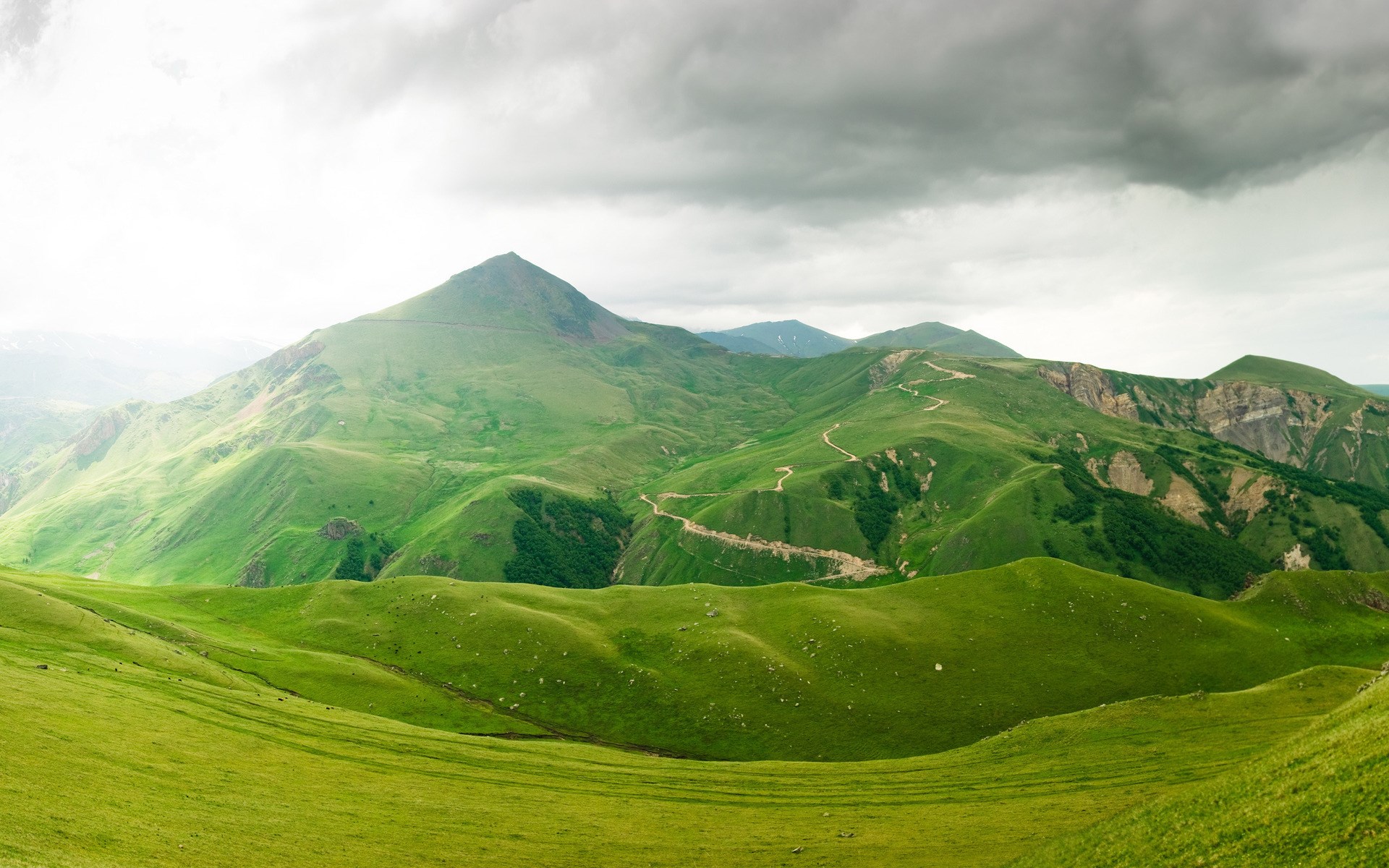 Green rolling hills and mountains under a cloudy sky HD Wallpaper