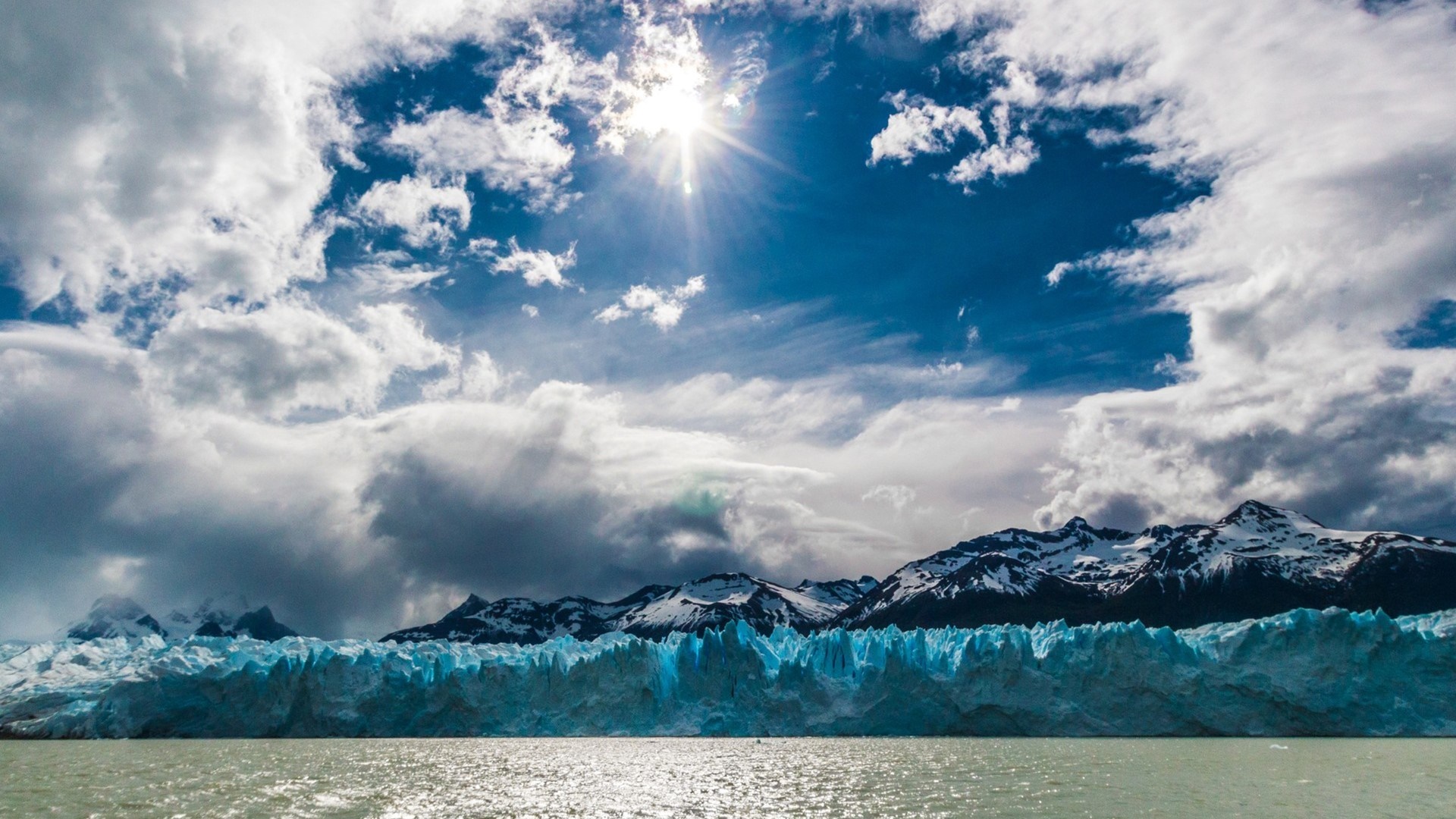 Glacier and snow-capped mountains under a cloudy sky with sun HD Wallpaper