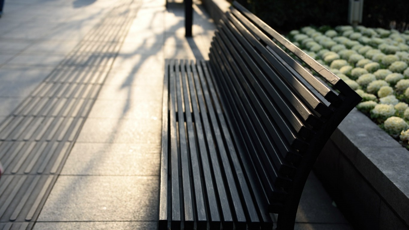 A dark slatted bench on a sidewalk with plants HD Wallpaper