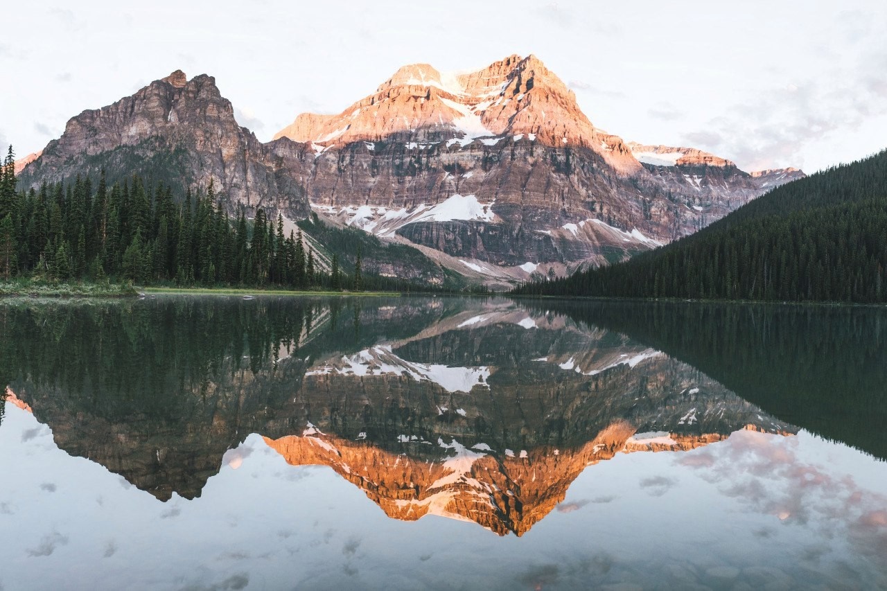 Mountain range reflected in a calm lake with pine forest HD Wallpaper