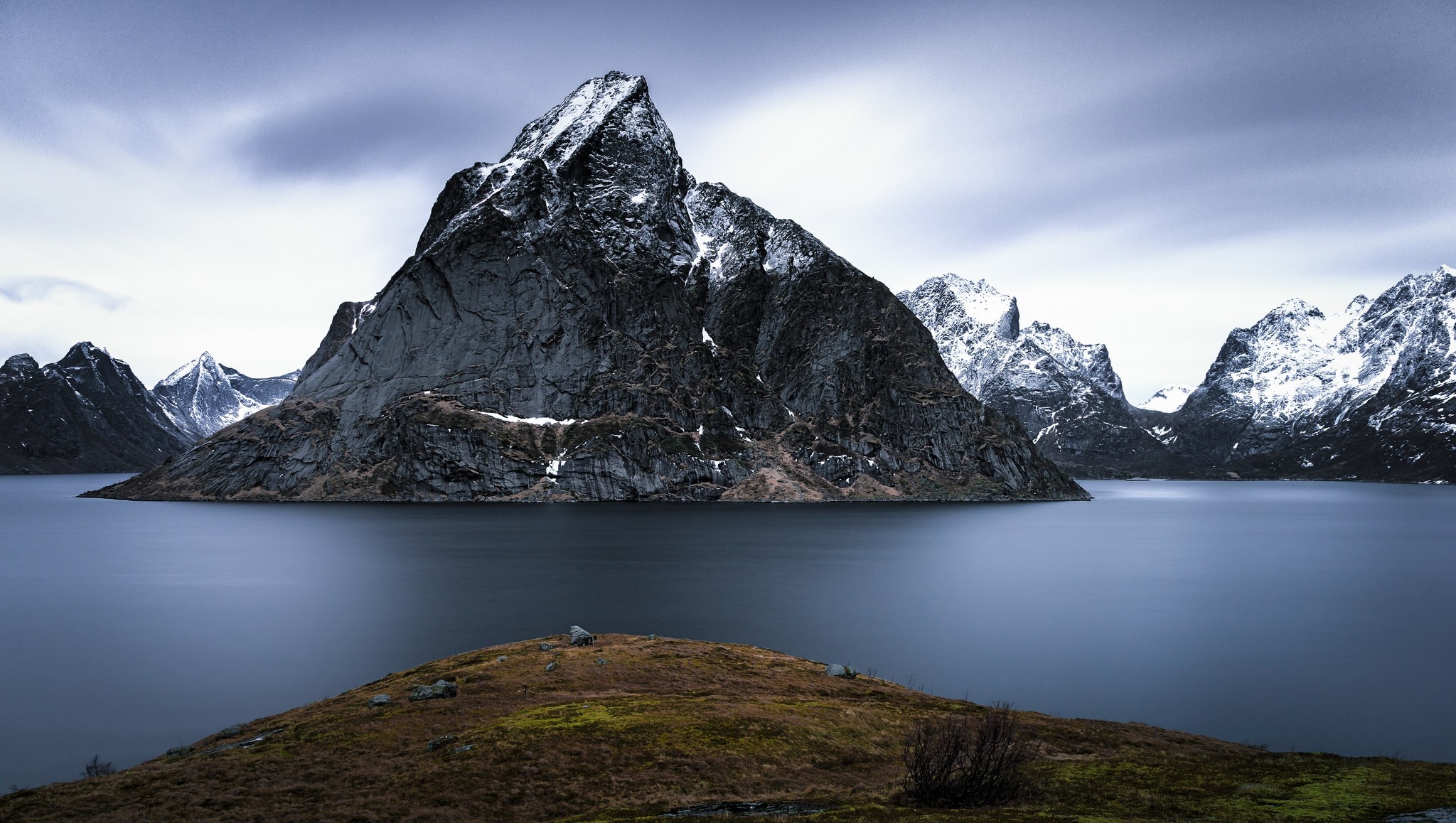 Snow-capped mountains rise from a calm lake under a cloudy sky HD Wallpaper