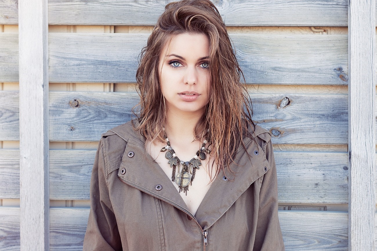 Woman with blue eyes and necklace in front of wooden wall HD Wallpaper