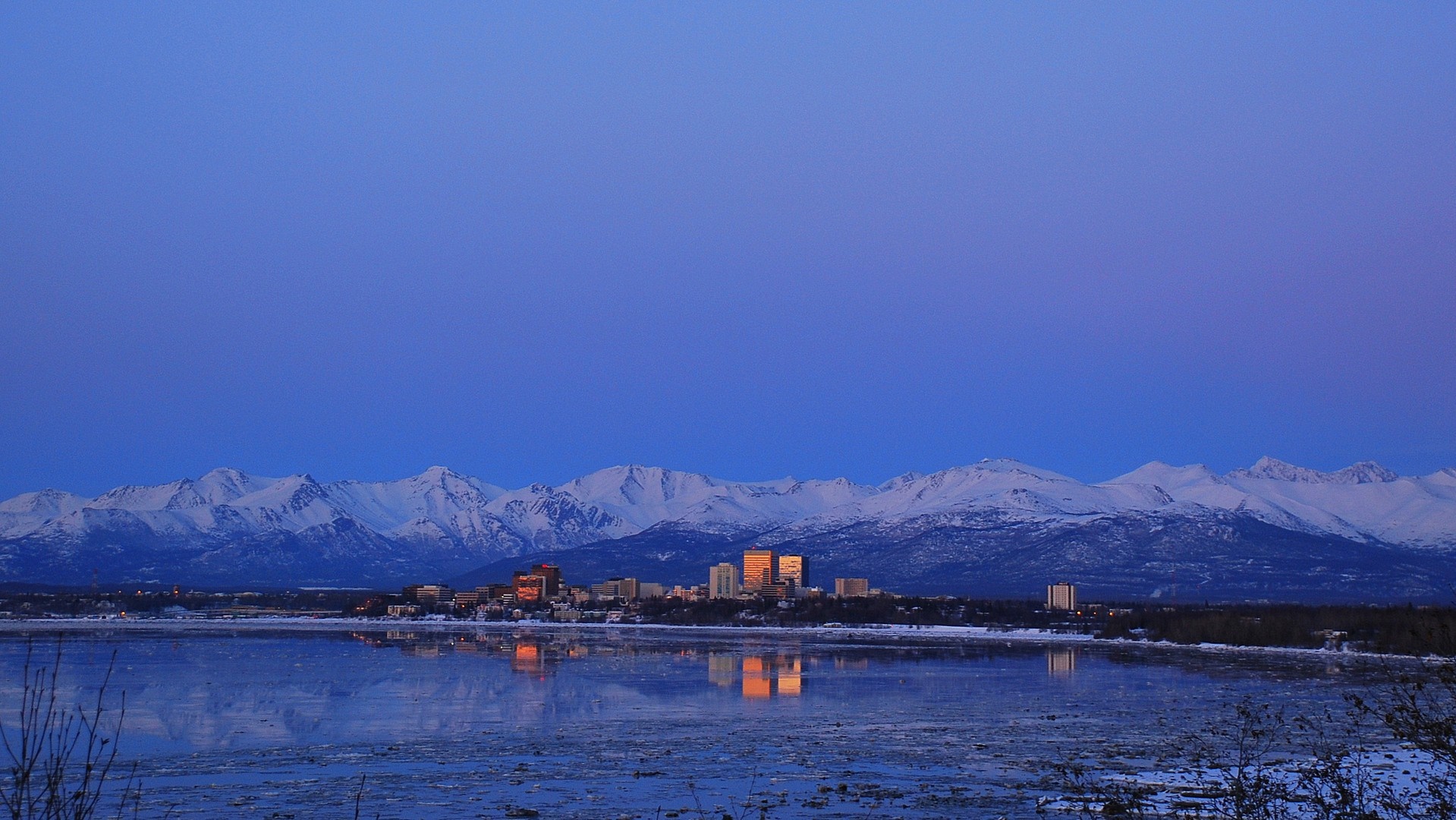Cityscape reflected in water with snow-capped mountains at dawn HD Wallpaper
