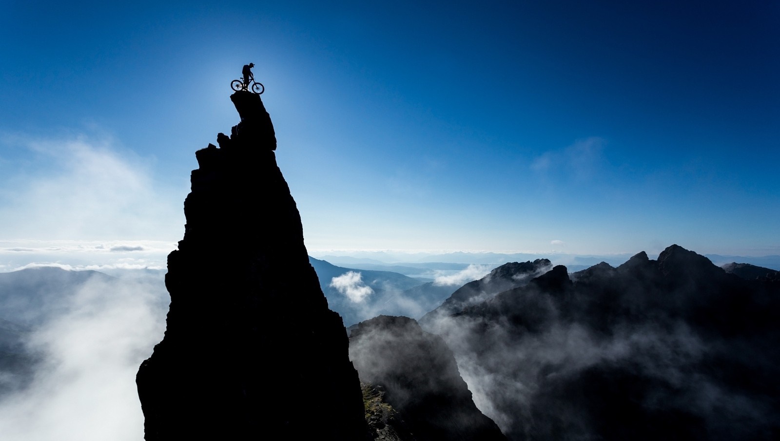 Mountain biker silhouetted on a rocky summit against a bright blue sky HD Wallpaper