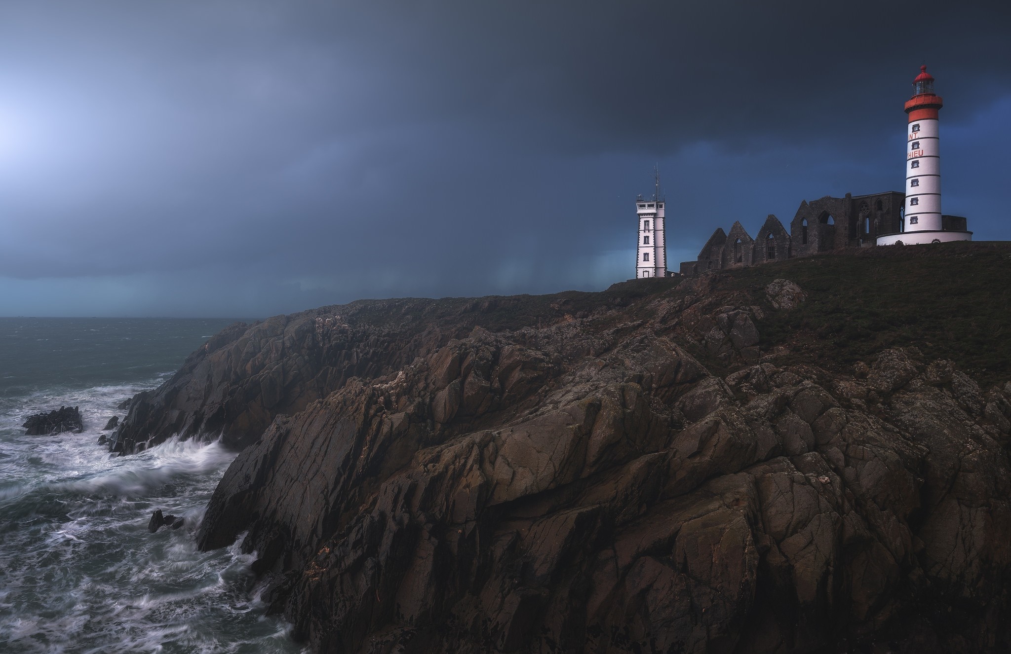 Lighthouse and ruins on a rocky coast during a storm HD Wallpaper
