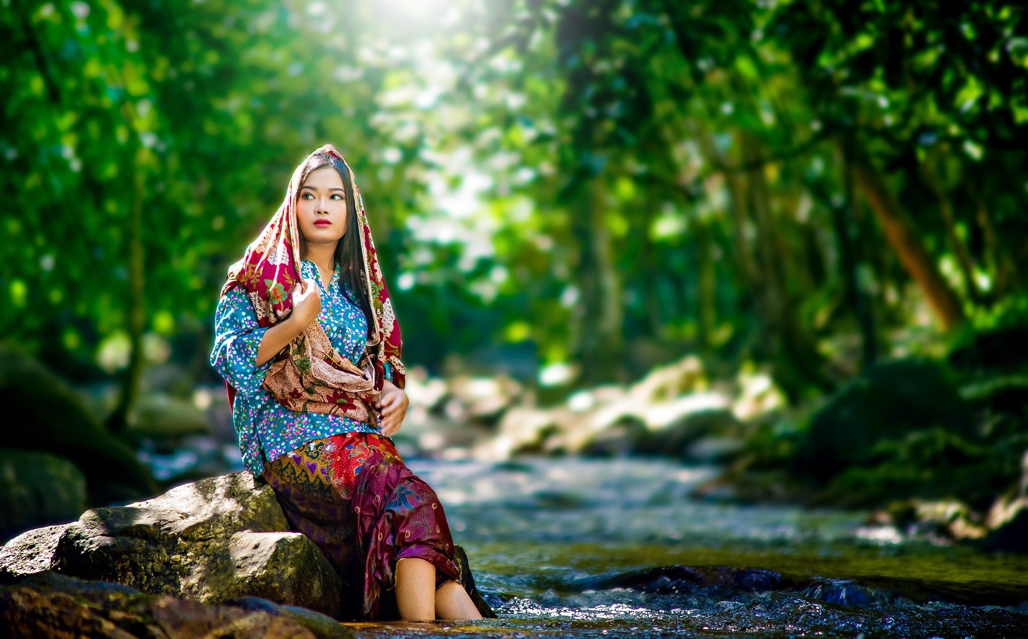 Woman in traditional clothing sitting by a stream in a forest HD Wallpaper