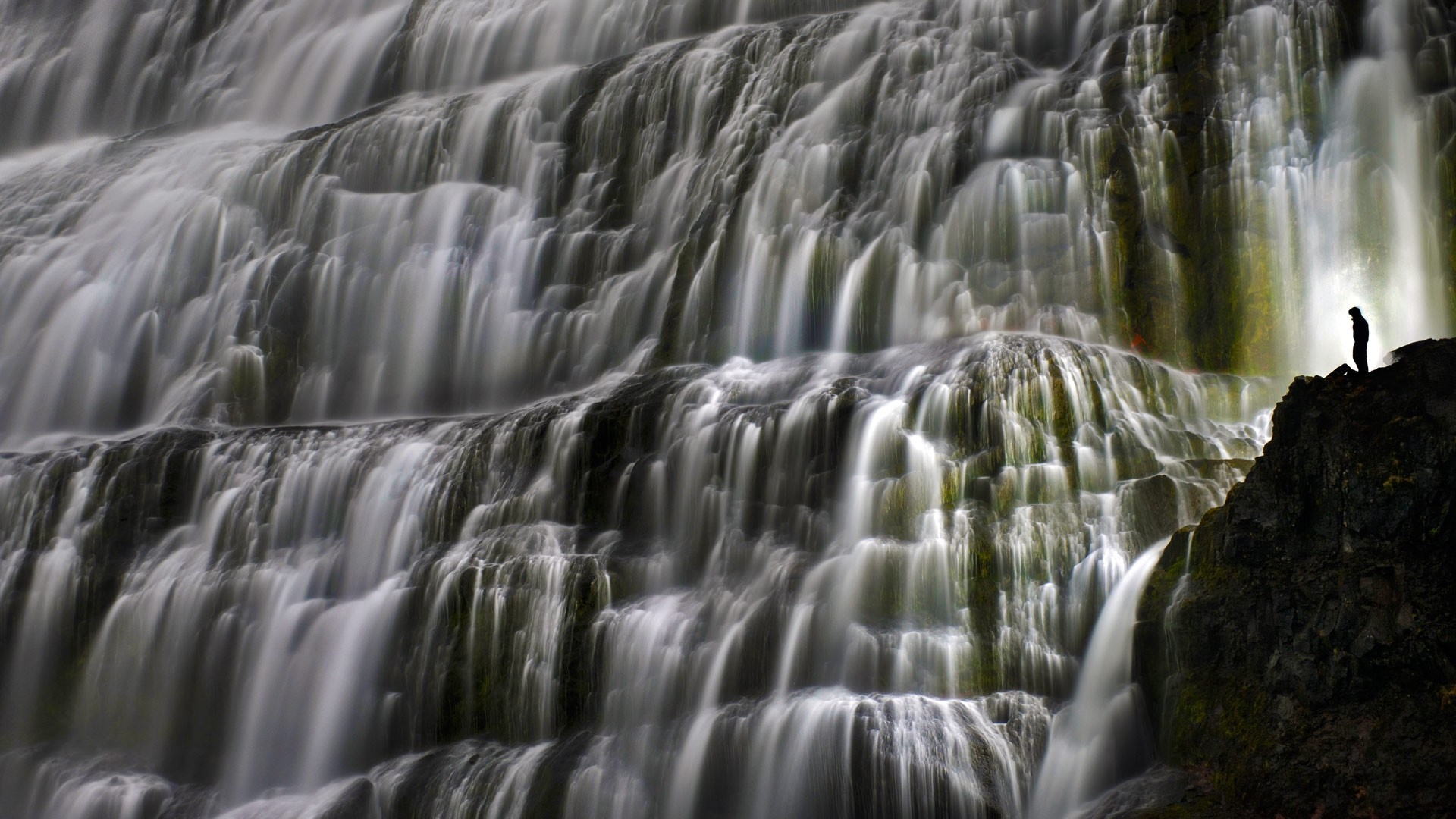 Silhouette of a person standing on a rock next to a cascading waterfall HD Wallpaper