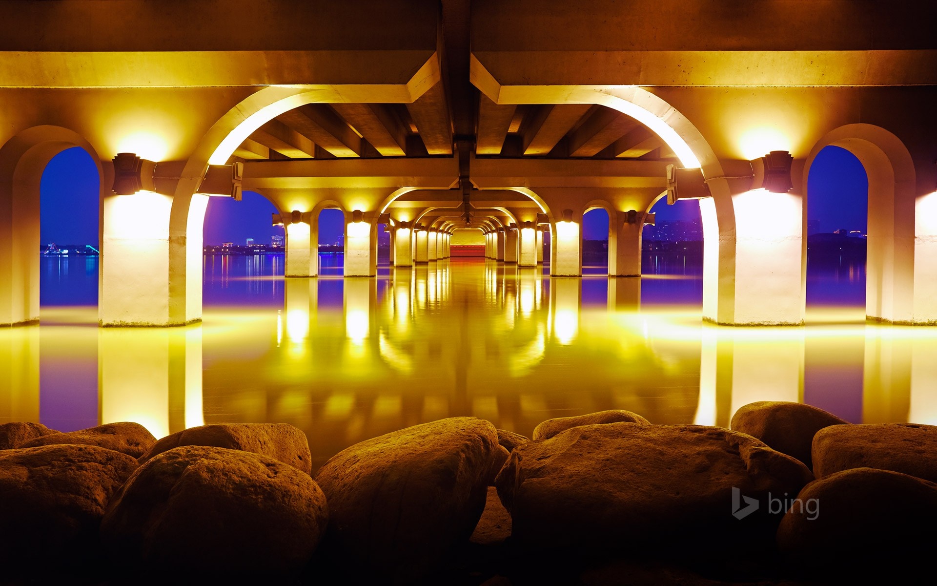 Bridge arches illuminated at night over water with rocks HD Wallpaper