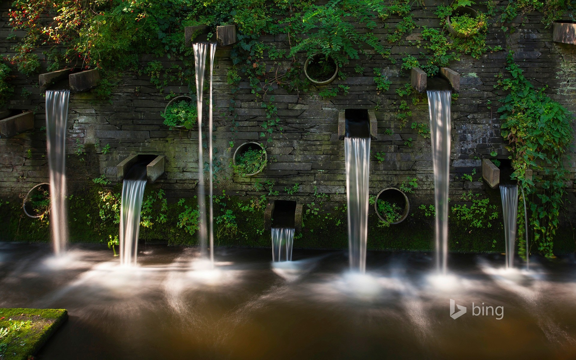 Waterfalls cascading down a stone wall into a stream HD Wallpaper