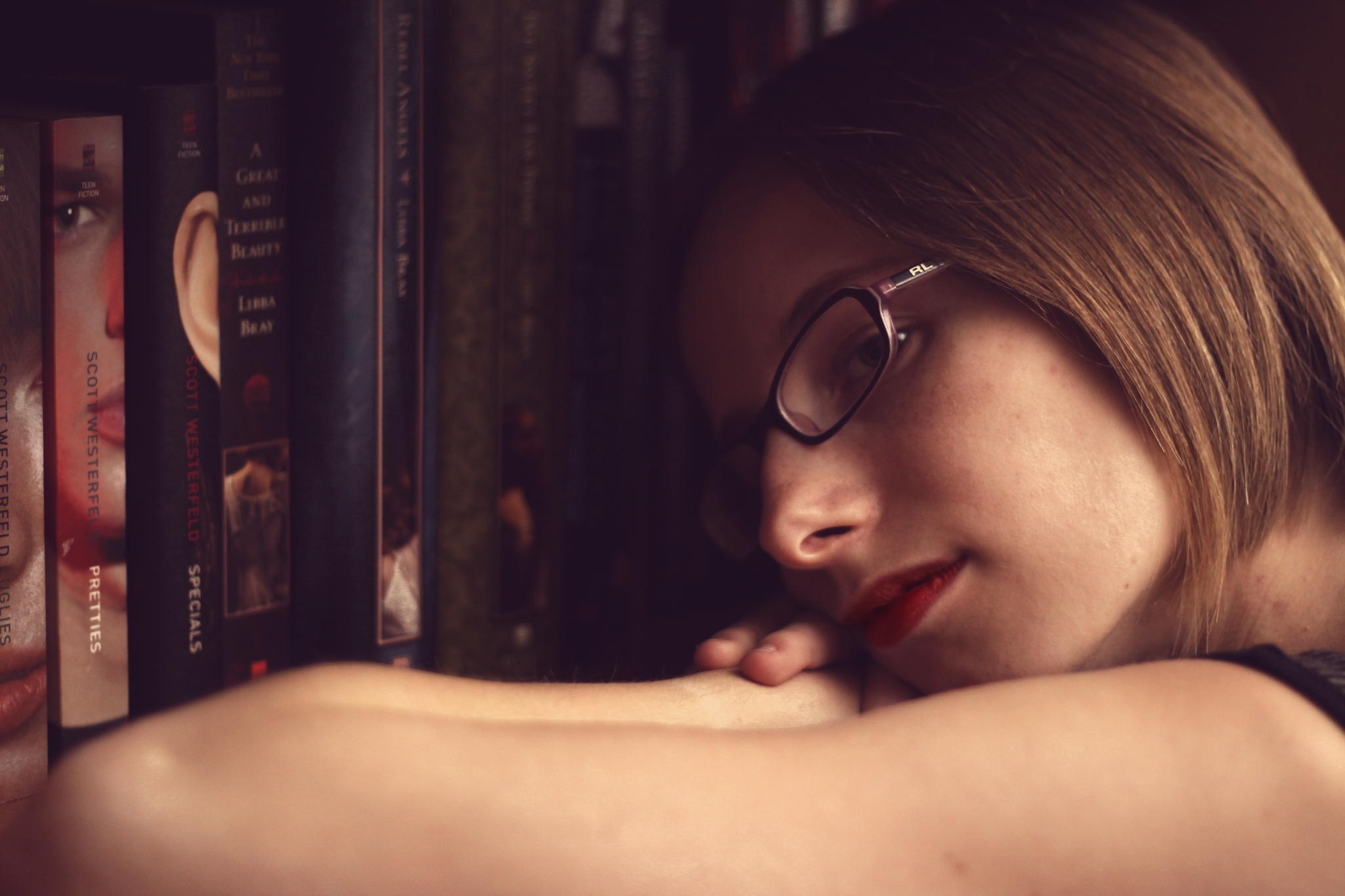 Woman with glasses resting head on arms in front of bookshelf HD Wallpaper