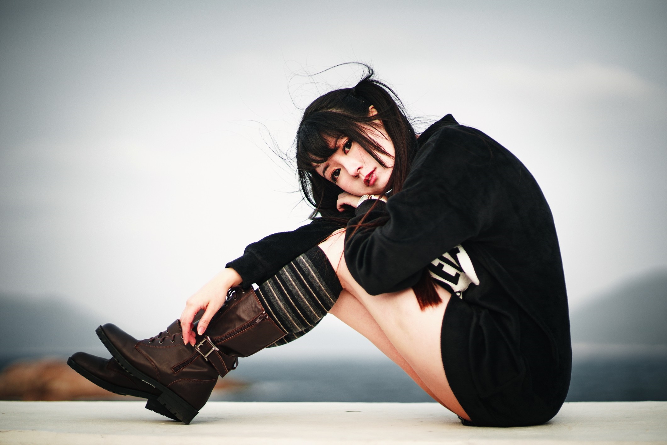 Asian woman wearing black hoodie and boots sitting outdoors HD Wallpaper