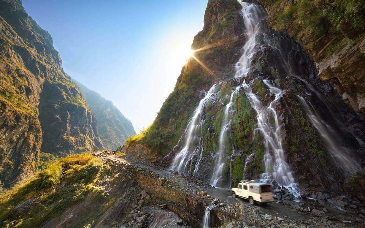 Off-road vehicle driving past a waterfall on a mountain road in Nepal HD Wallpaper