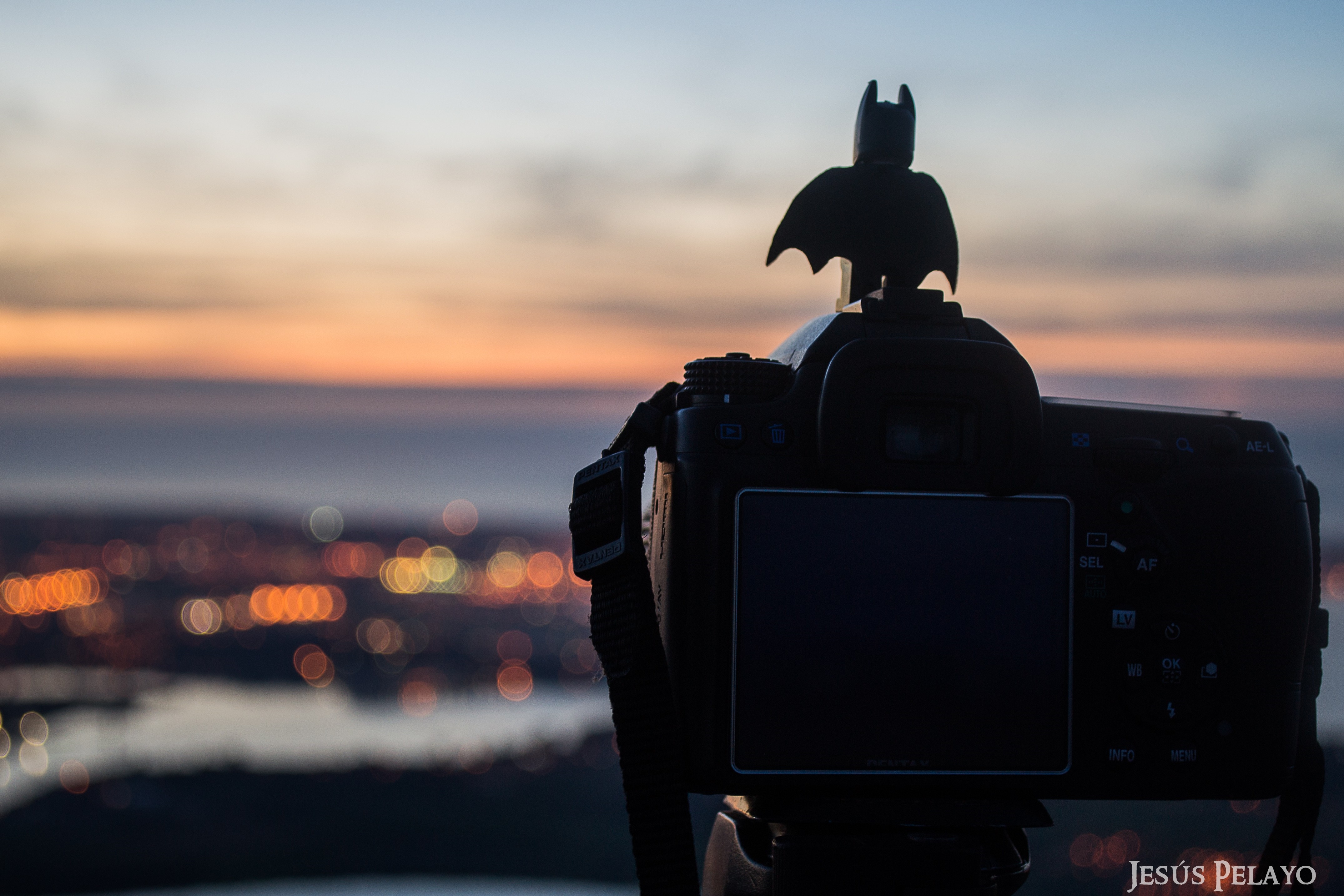 Batman figurine on camera overlooking city at sunset HD Wallpaper