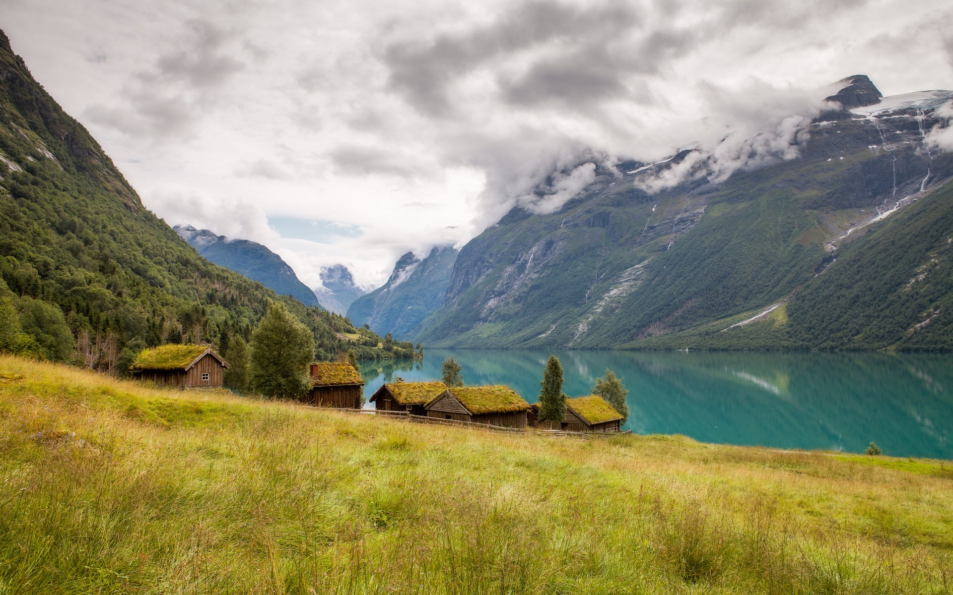Wooden cabins with grass roofs by a lake in the mountains HD Wallpaper
