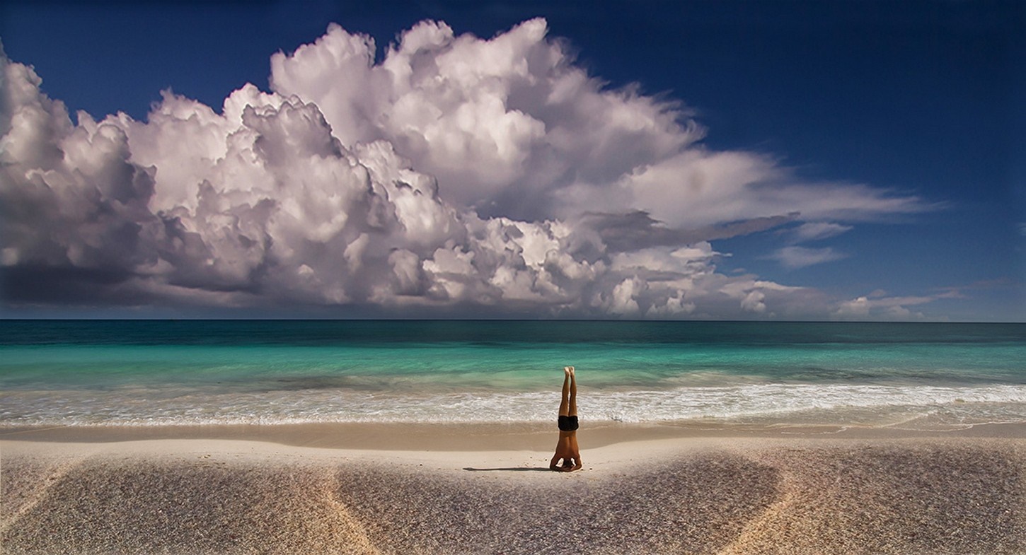 Man doing a headstand on a beach with dramatic clouds HD Wallpaper