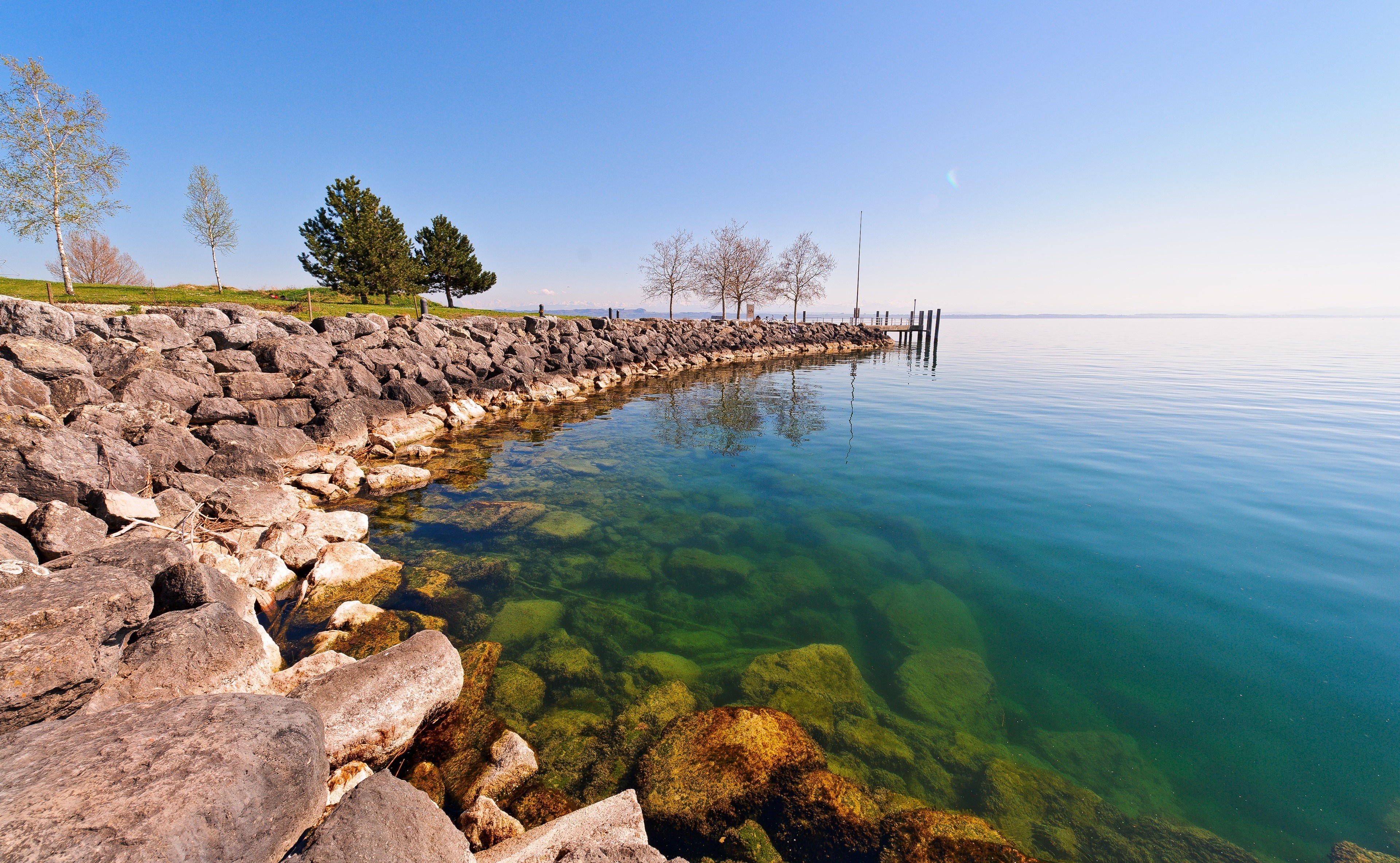 Rocky shore of a clear lake with a wooden pier HD Wallpaper