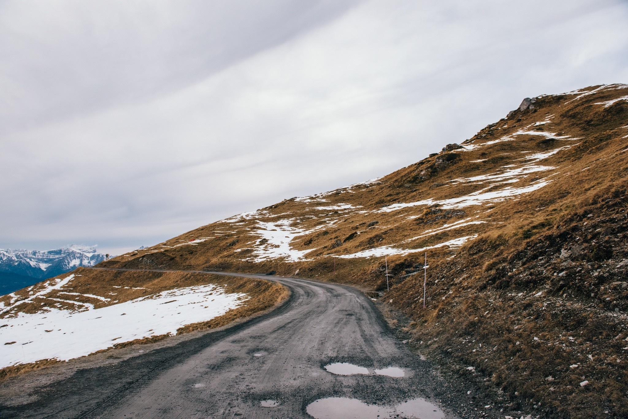 Winding mountain road with snow patches and distant mountains HD Wallpaper