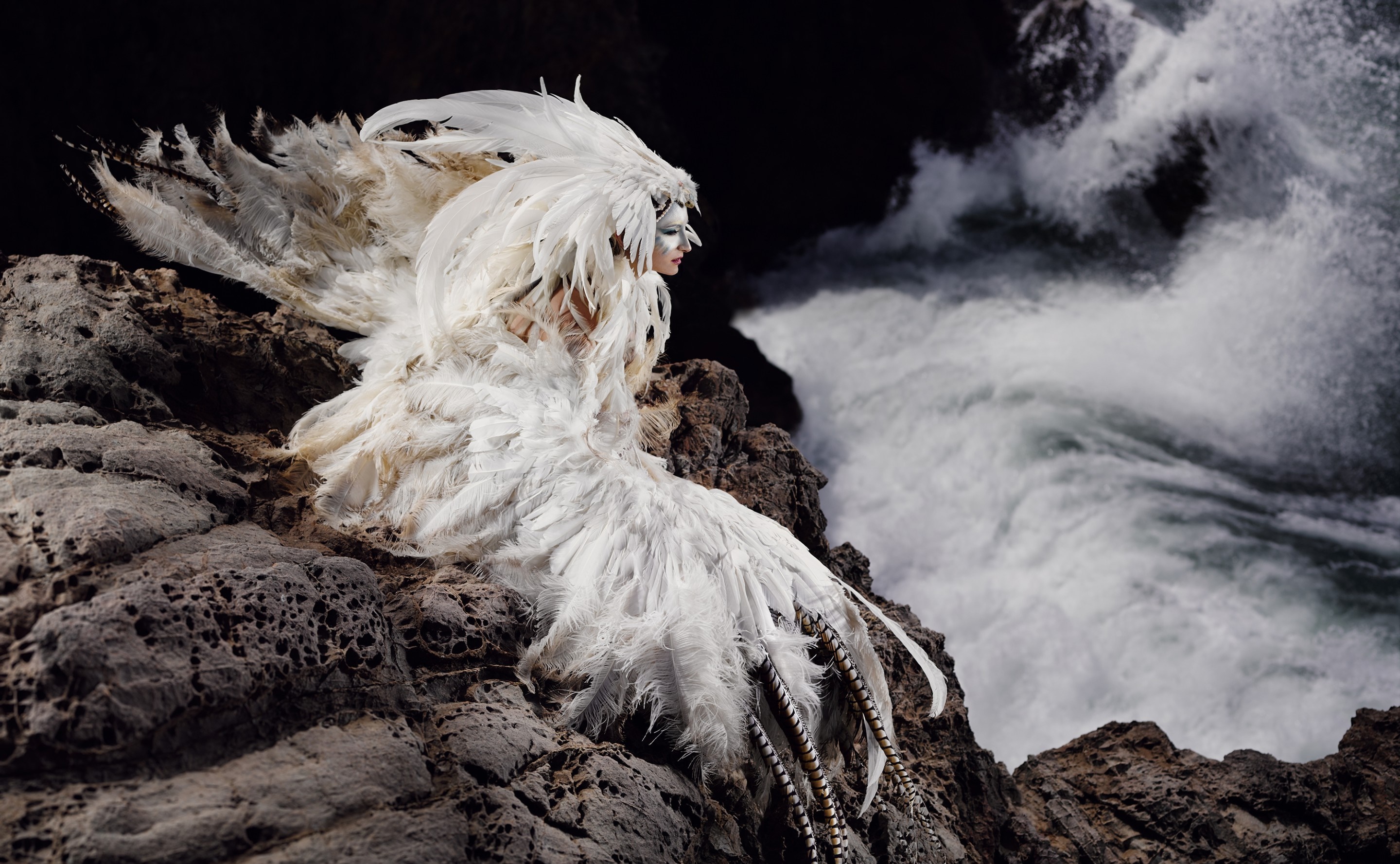 Woman in white feathered costume on rocks by waves HD Wallpaper