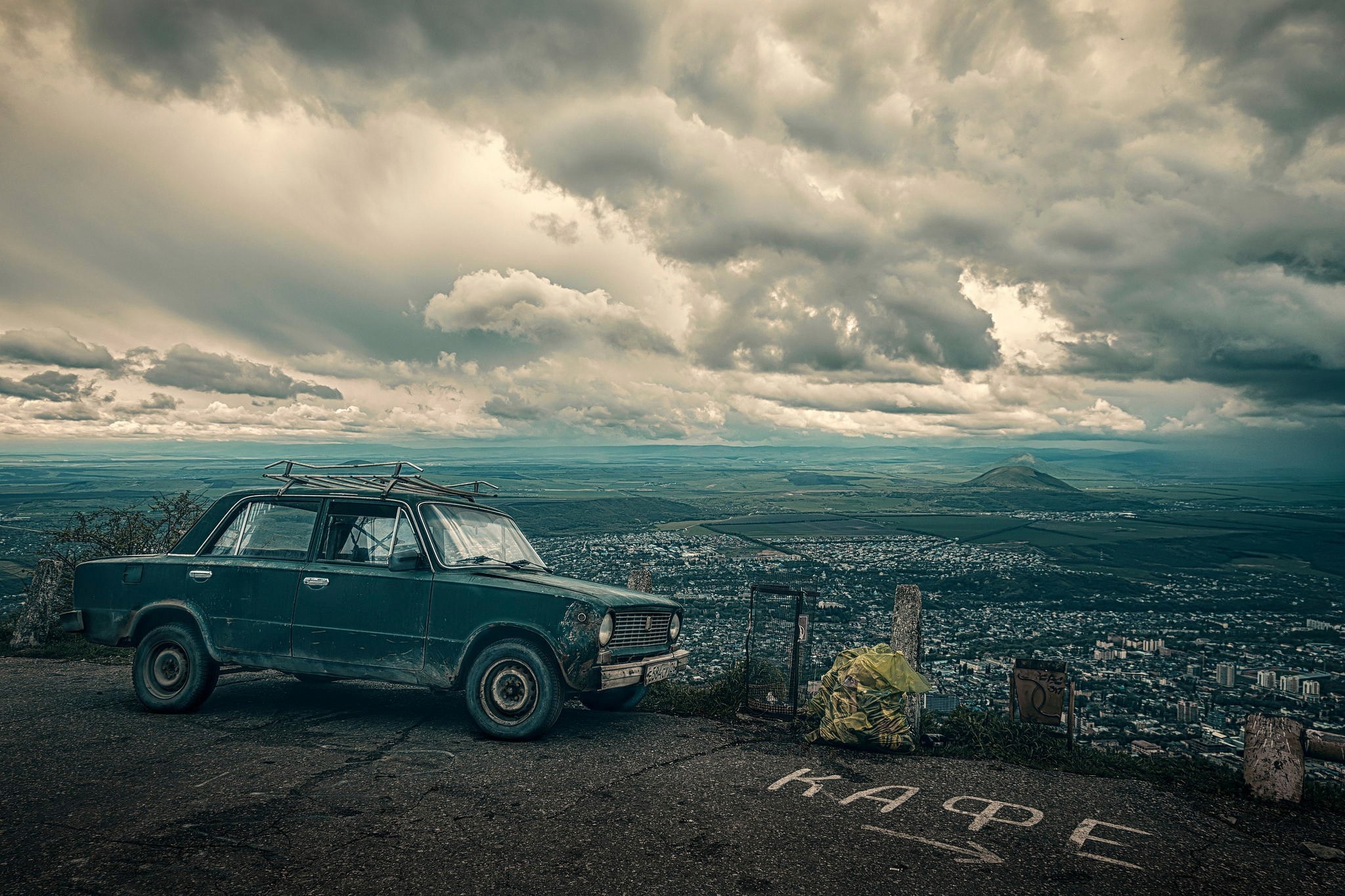 Old car parked on a cliff overlooking a city and mountains HD Wallpaper