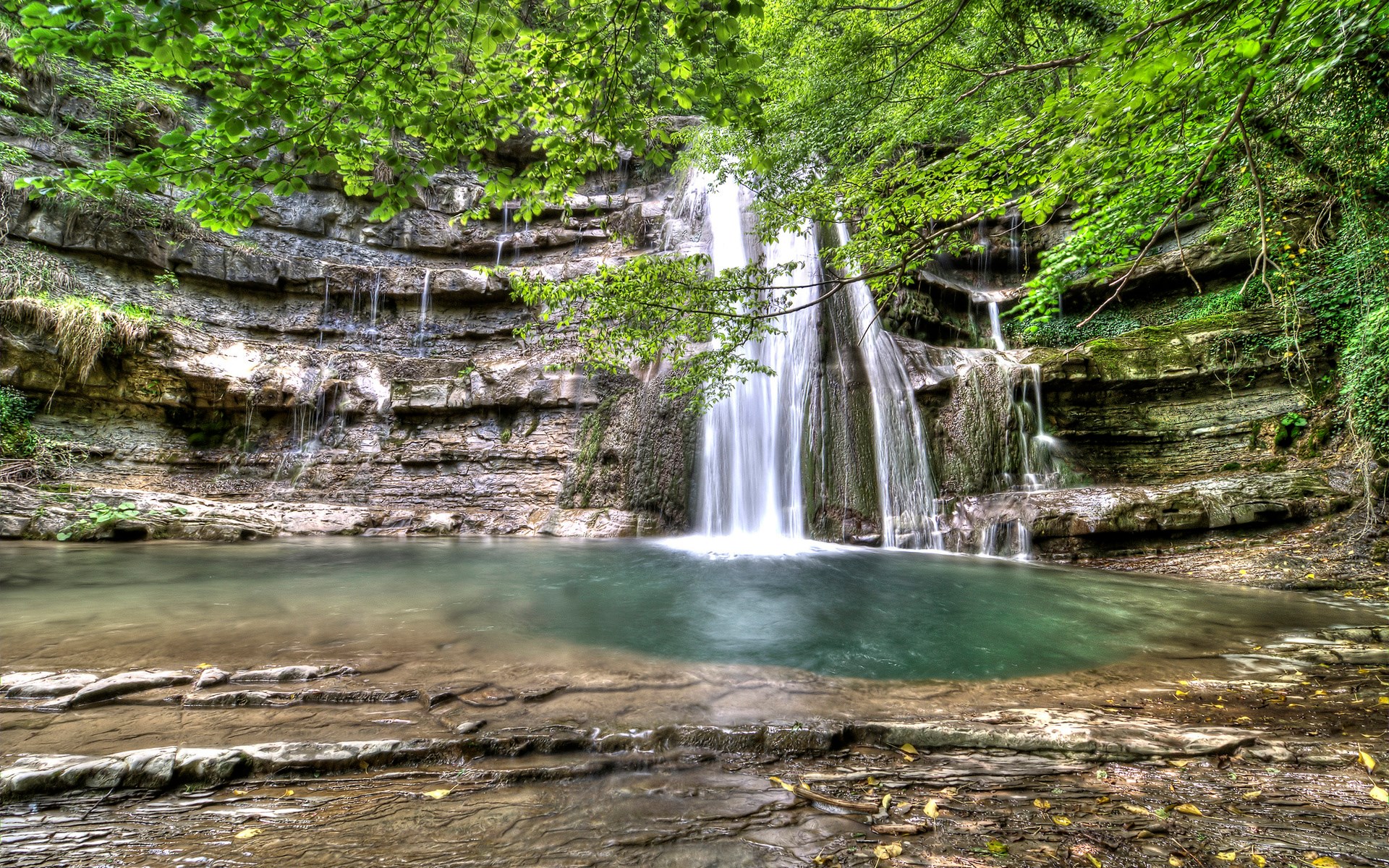 Waterfall cascading into a clear pool surrounded by rocks and trees HD Wallpaper