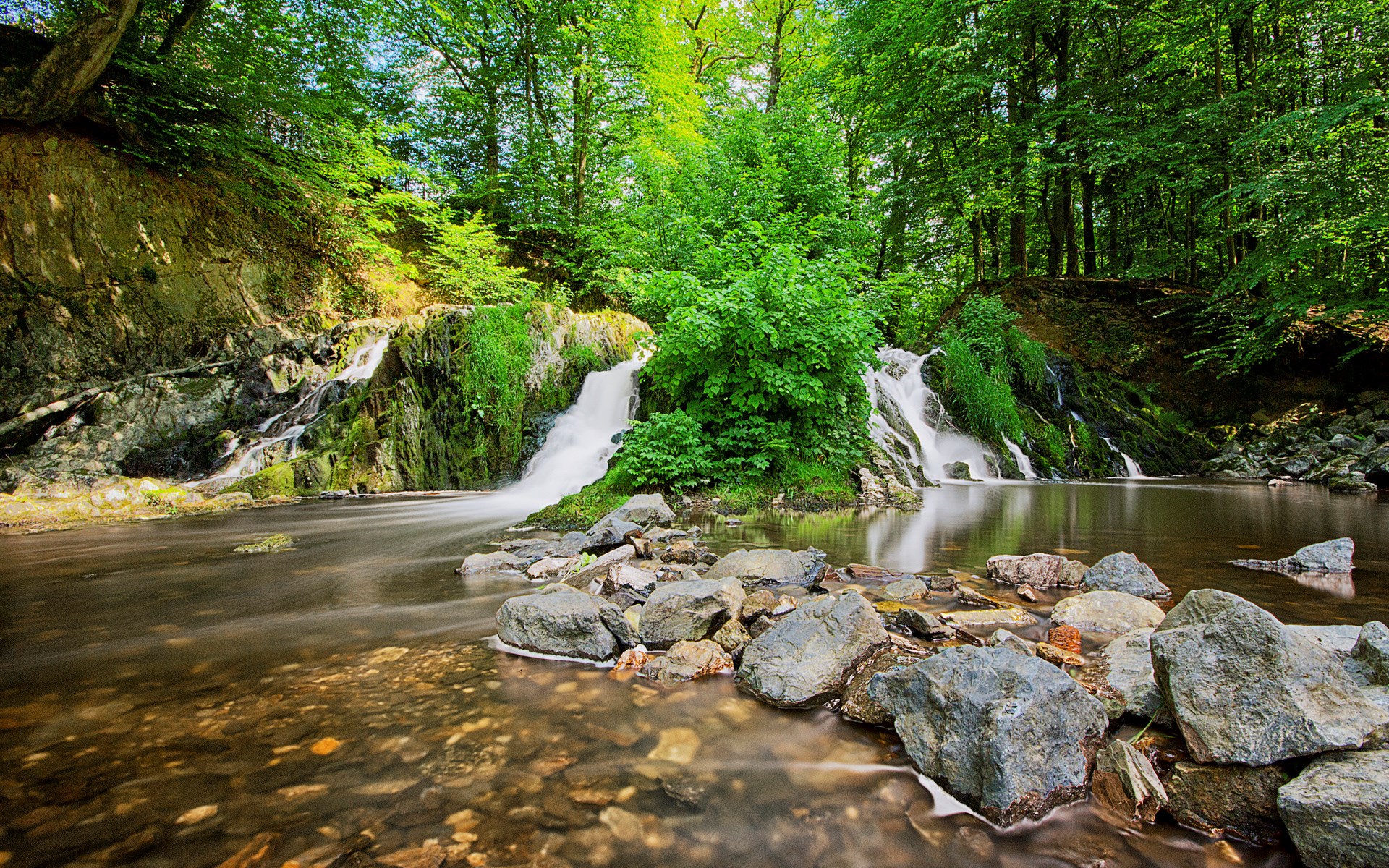 Waterfalls in a lush green forest with rocks in the foreground HD Wallpaper