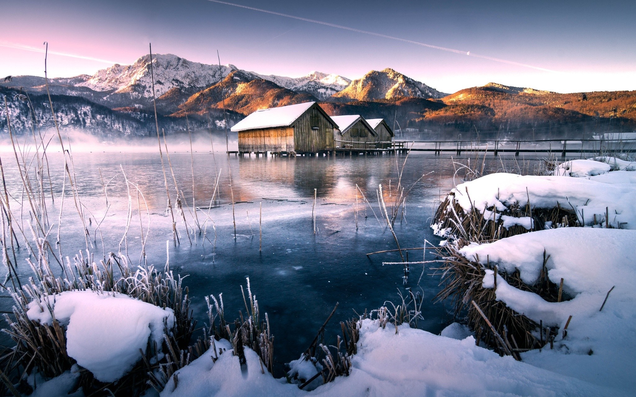Wooden boathouses on a frozen lake with snow-capped mountains HD Wallpaper