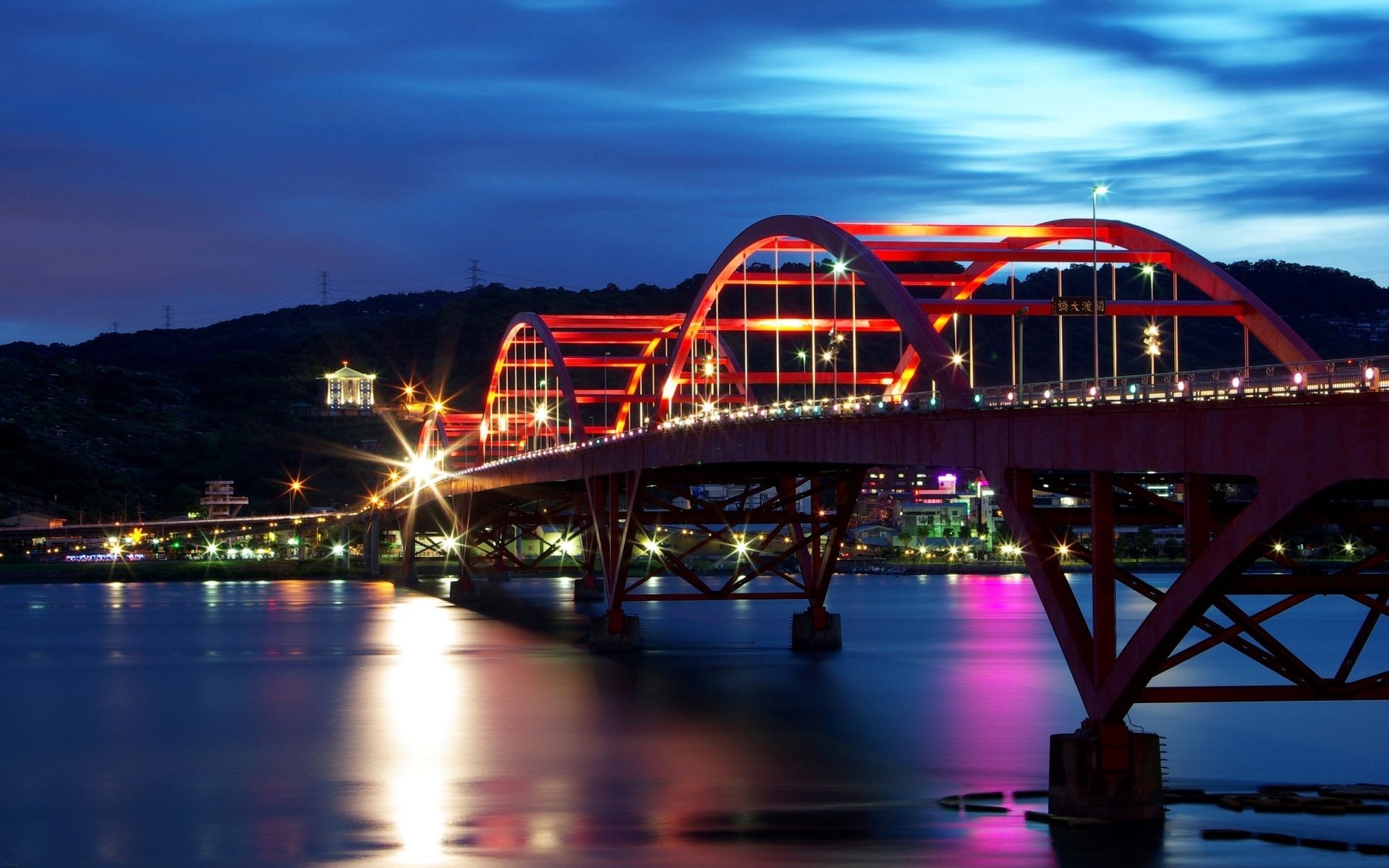 Red arched bridge over a river at night with city lights HD Wallpaper