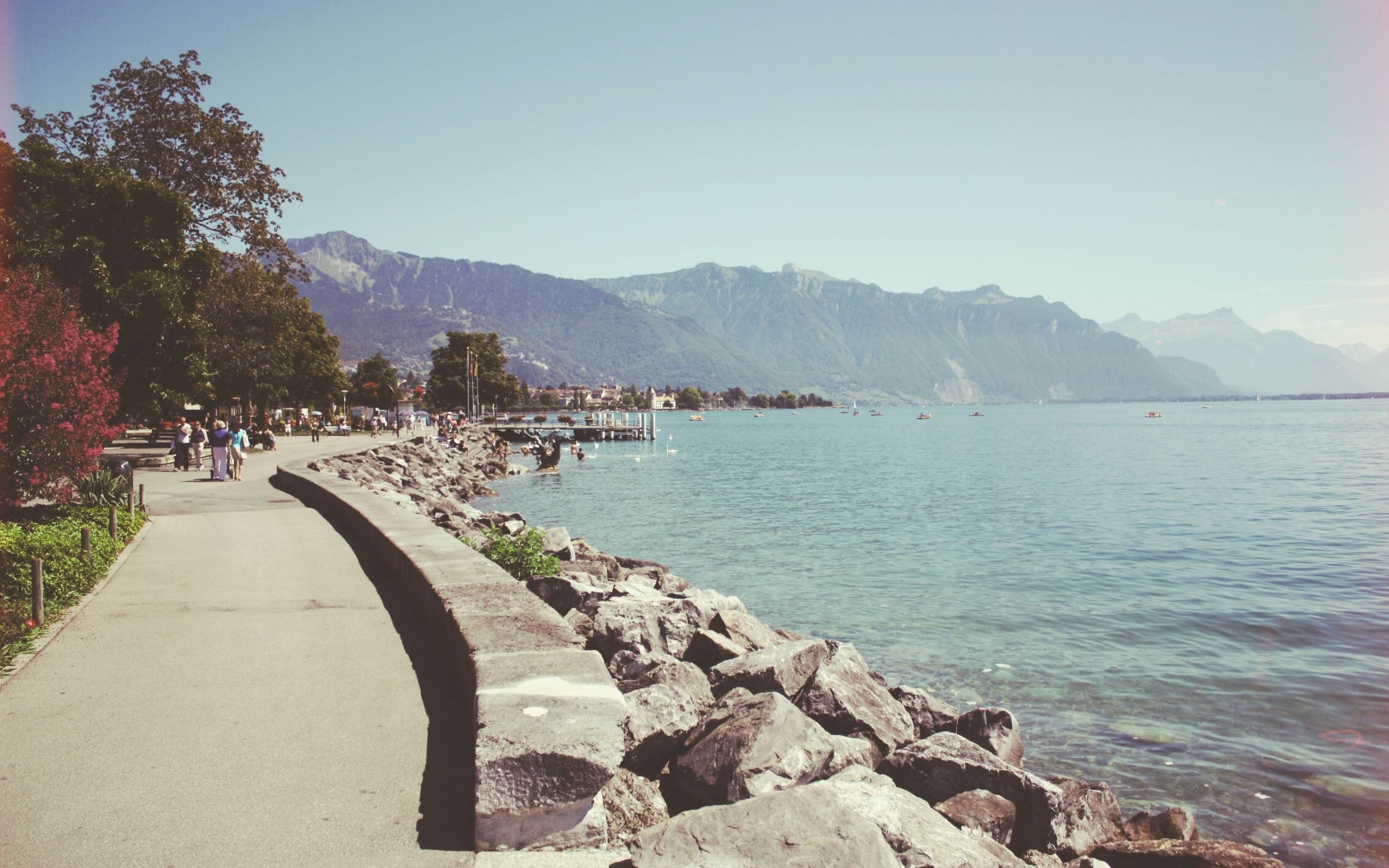 People walking along a paved path by a rocky shore and a lake with mountains in the background HD Wallpaper