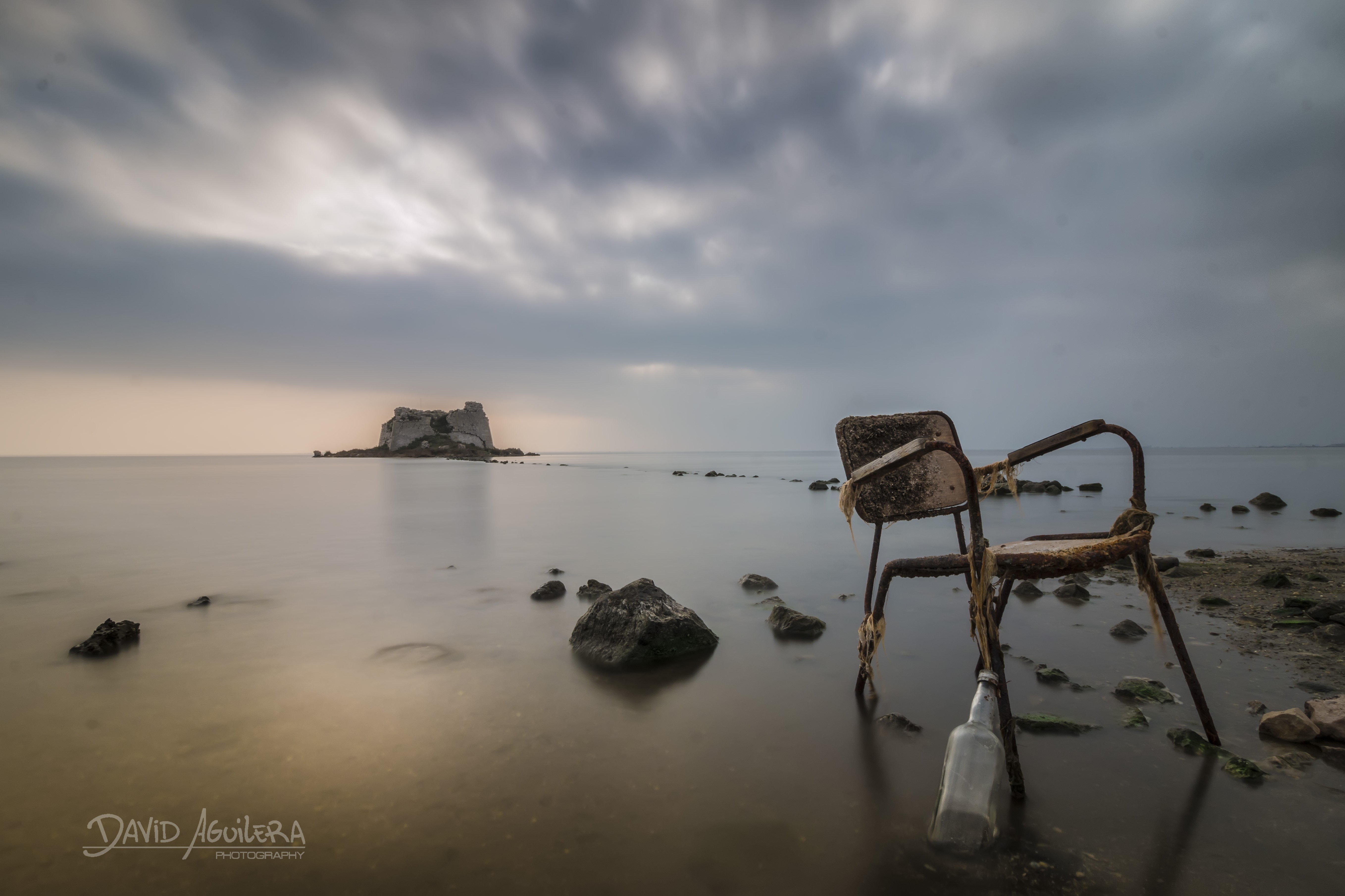 Rusty chair and bottle on a rocky shore with island in the sea HD Wallpaper