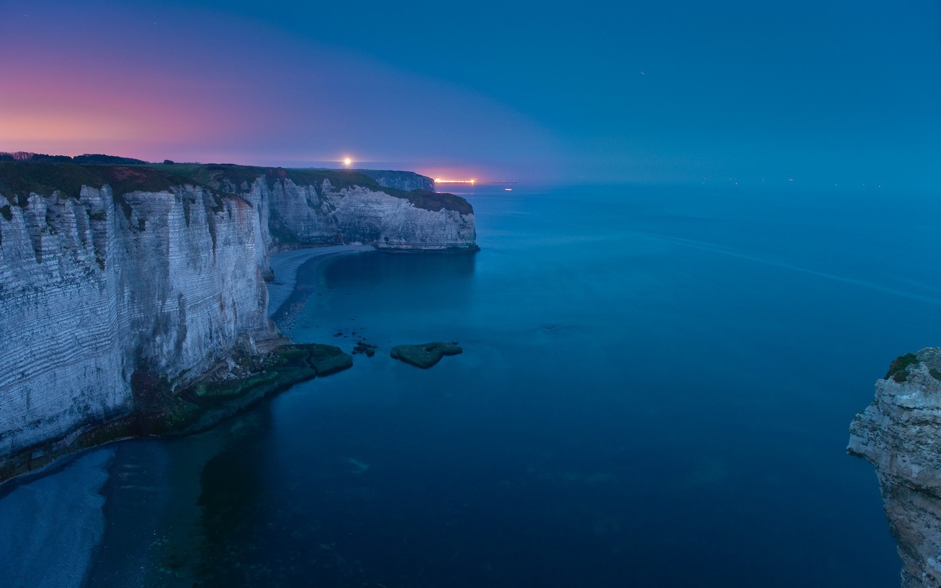 Cliffs and ocean at dusk with distant lights HD Wallpaper
