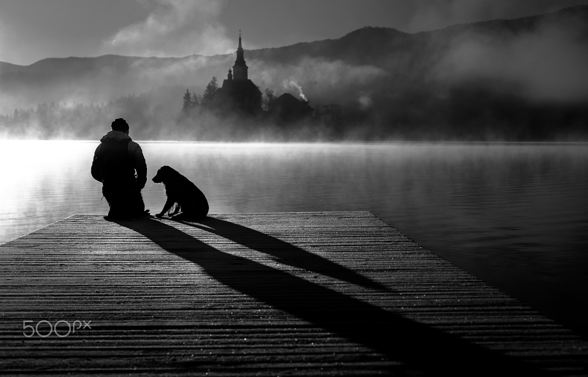Man and dog on a pier with a misty lake and church HD Wallpaper